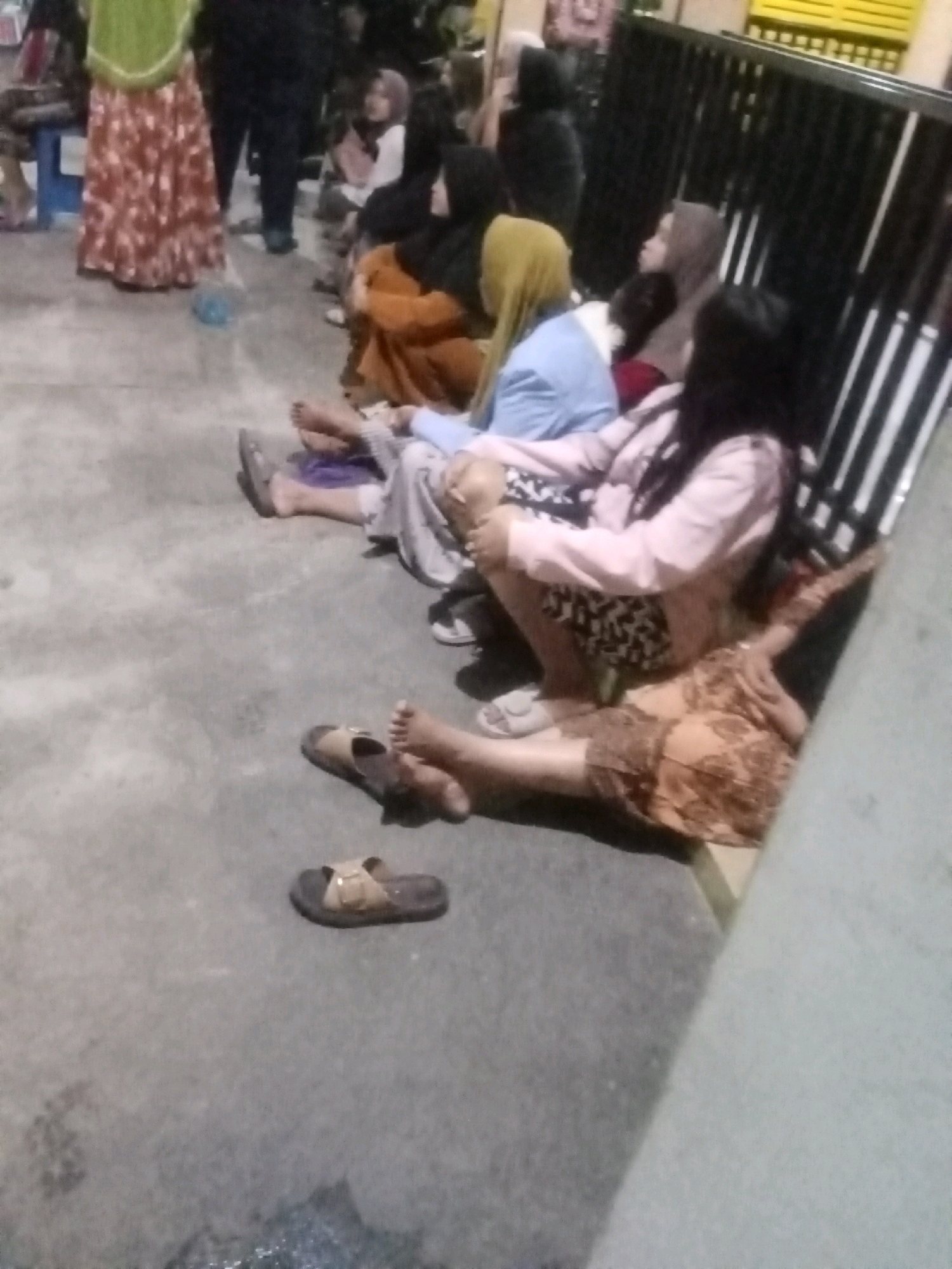 In Keplaksari, Indonesia, a group of women are seated on the ground, appearing to be waiting or resting. The scene is outdoors, with a concrete ground surface. Several women are wearing traditional Indonesian attire, including headscarves and patterned skirts. Some are barefoot, while others wear sandals. One woman in the foreground is lying down on her side, wearing a patterned dress. Another woman is seated next to her, wearing a pink jacket and patterned shorts. In the background, a black metal fence with horizontal bars is visible, and beyond that, a yellow structure, possibly part of a building. A red and white patterned skirt is also visible on a woman standing on the left side of the frame. The overall impression is of a casual gathering or waiting area.