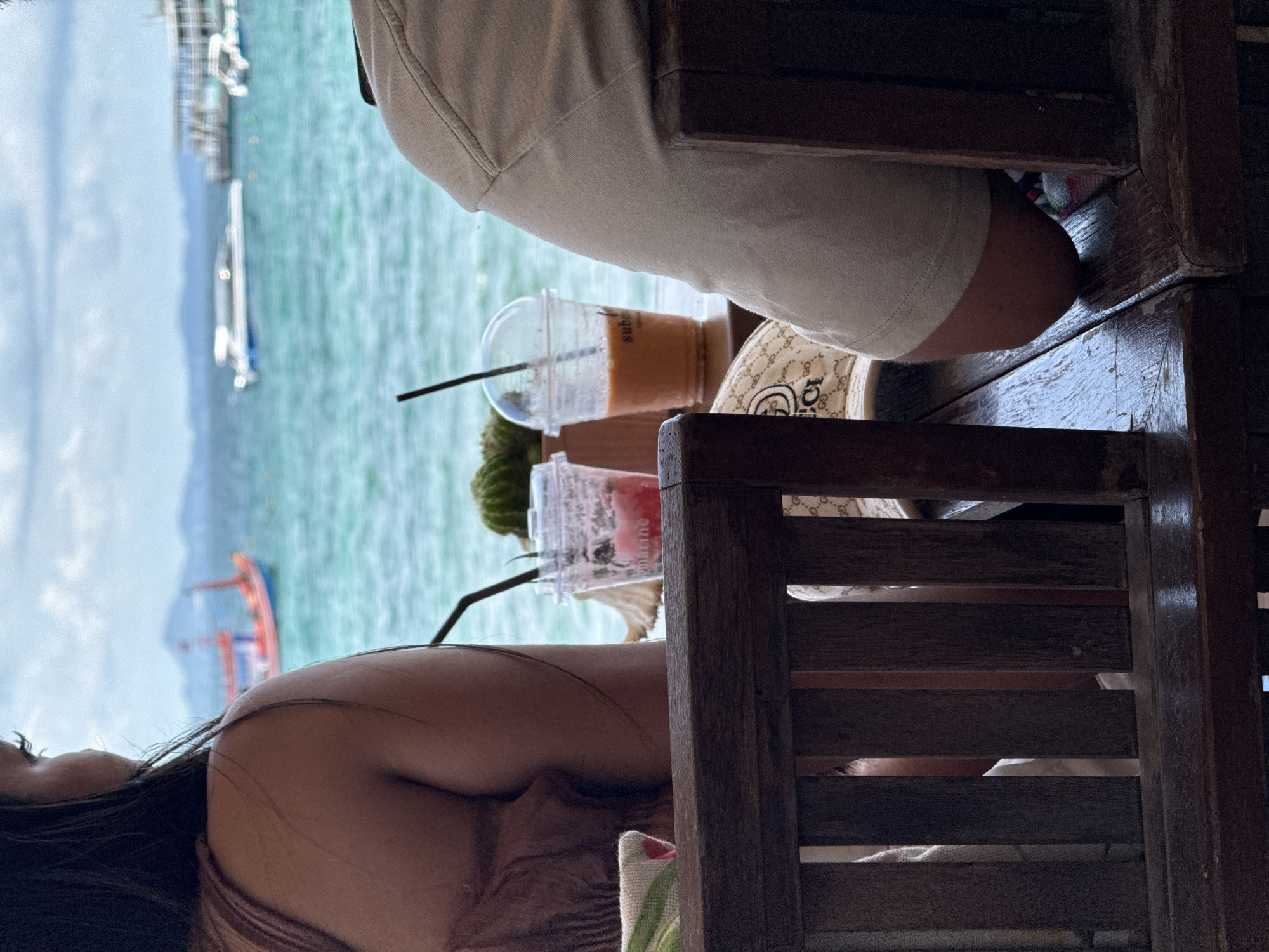 The image shows two individuals seated at a wooden table in what appears to be an outdoor setting with a view of the ocean. The location is identified as Pattaya City, Thailand, based on the presence of water and boats in the background.
The first individual is wearing a light-colored top. They are sitting on a dark wooden chair.
The second individual, seated to the left, has long dark hair and is wearing a brown top. They are also sitting on a wooden chair.
On the table between them are two drinks in clear plastic cups with straws. One appears to be a Thai iced tea and the other is a red mixed drink. A Gucci wallet/handbag rests on the table as well.
In the background, there is an ocean with several boats. The sky is partially cloudy.