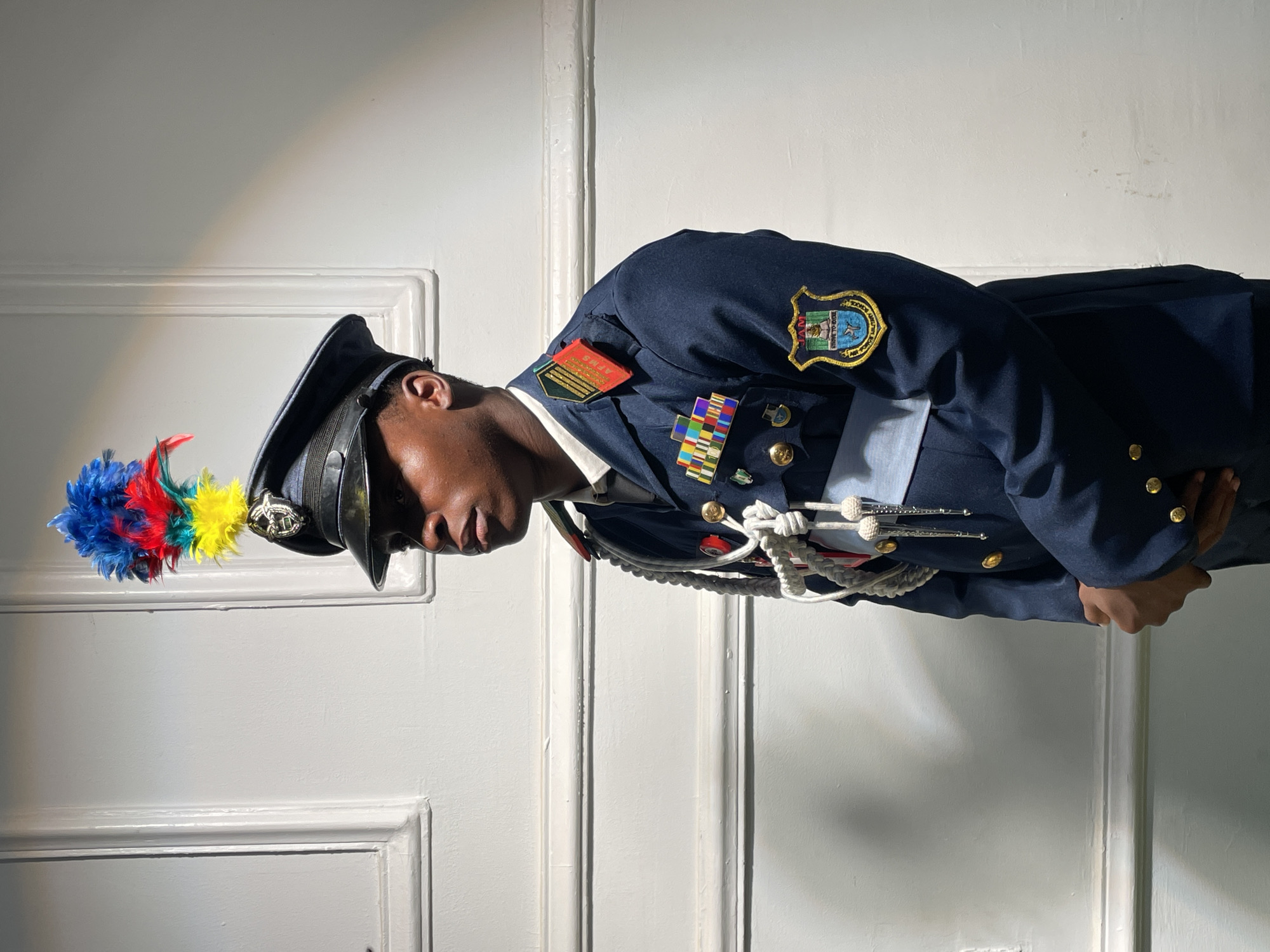 A person in a dark blue military-style uniform with gold buttons, a light blue sash, and various medals and insignia is standing indoors against a white paneled wall. The person is wearing a hat with a colorful plume, possibly red, yellow, and blue. The hat also features a silver emblem. The person's head is slightly bowed. The uniform has a patch on the shoulder featuring an emblem with text and imagery. The environment suggests an official setting or event. No specific details about the city of Jos, Nigeria are discernible from the image.