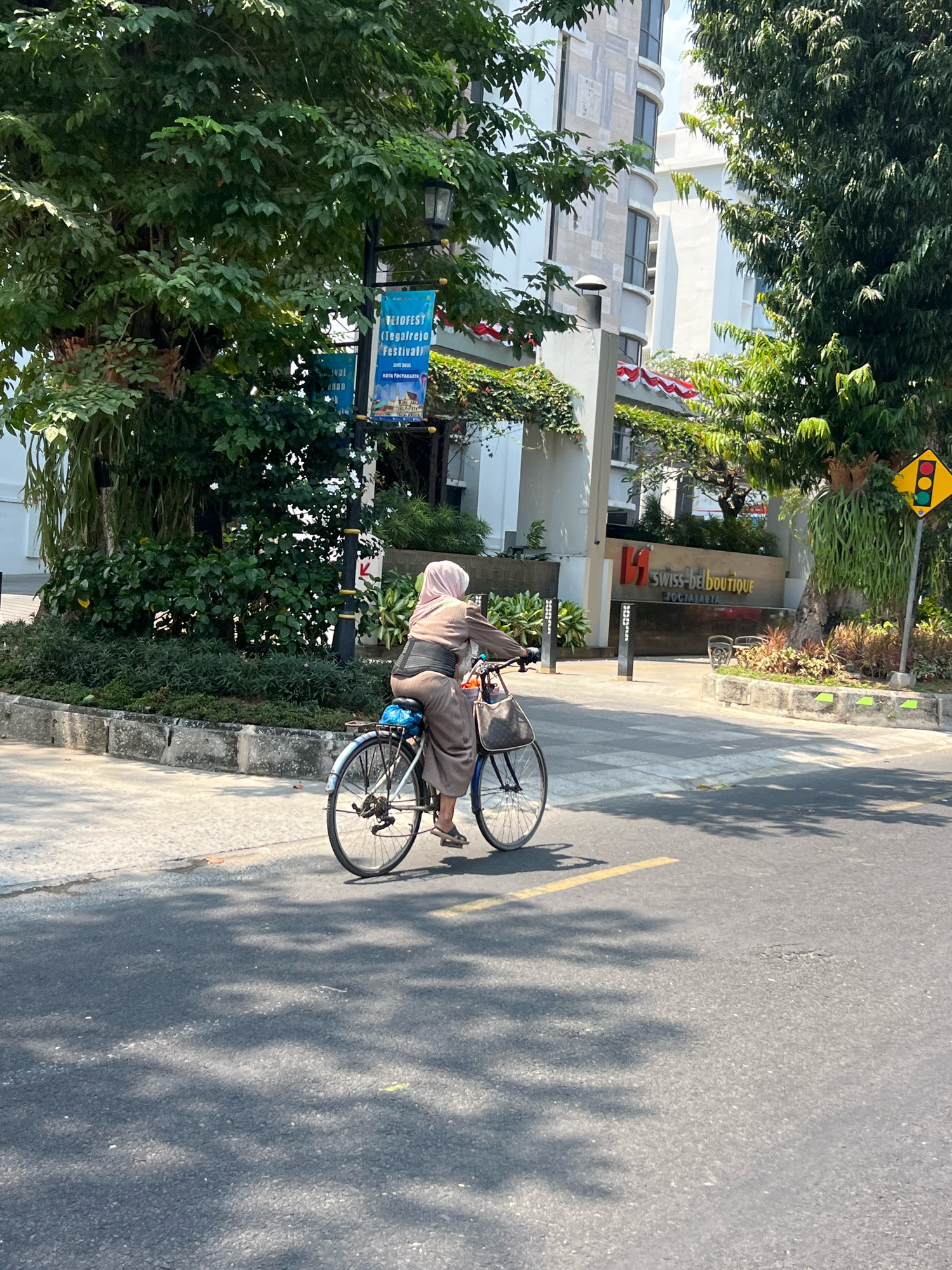 A woman wearing a hijab and a loose-fitting brown outfit rides a bicycle on a paved road in Yogyakarta, Indonesia. The bicycle is dark colored with a blue seat cover and a basket attached to the front. The woman is facing away from the camera, and her feet are resting on the pedals. She appears to be wearing sandals. The road has a yellow line marking its center. To the left of the road is a sidewalk with a stone border and green foliage. Behind the foliage, a light-colored building with multiple windows is visible. A street lamp with a banner is also present, and the banner has text on it that appears to be in Indonesian. To the right of the road, there is a large tree with lush green leaves. Further to the right, a yellow and red traffic signal is mounted on a pole. A sign with the logo "11 Swiss-bel boutique" and the word "YOGYAKARTA" is visible on another building in the background. The scene is brightly lit by sunlight, casting shadows from the trees onto the road.