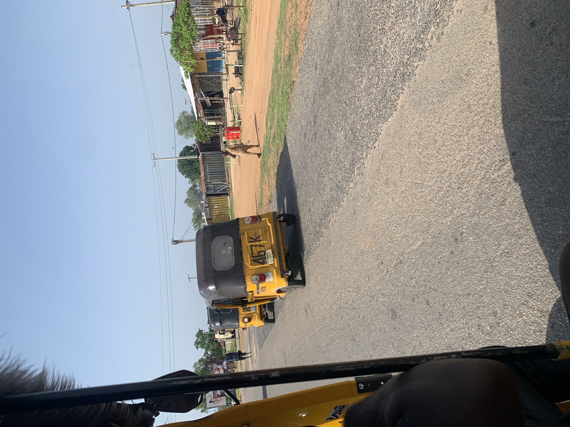 In this outdoor, daytime shot, the camera captures a street scene from within a yellow auto-rickshaw. The perspective is looking forward and slightly to the right. In the immediate foreground, the edge of the auto-rickshaw's interior, a black padded seat, and part of a metal support pole are visible on the left and bottom. The road ahead is paved and appears to be in good condition, with a distinct lighter-colored patch of gravel or sand on the right side of the lane.
Further down the road, two yellow auto-rickshaws are parked side-by-side. The one closer to the camera has a license plate that reads "467K" and appears to be a Ghanaian plate. The auto-rickshaw behind it is similar in color and design.
To the right of the road, a person wearing a light brown garment walks away from the camera, carrying what looks like a stick. Behind them are a row of small structures, possibly market stalls or simple buildings with corrugated metal roofs. A tree with green foliage is visible near the right edge of the frame. The environment appears somewhat rural or semi-urban, with utility poles and overhead wires present. The sky is clear and blue.