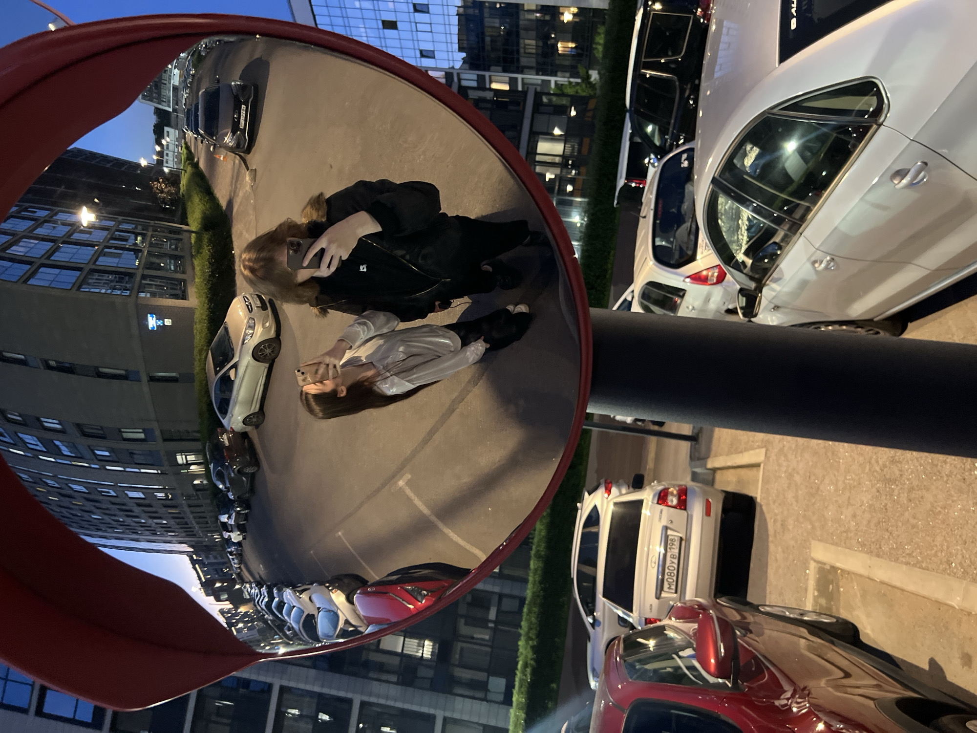 The image captures a late evening scene in Saint Petersburg, Russia, centered on a large convex traffic mirror with a red frame, mounted on a dark pole. Two young females are reflected in the mirror, both holding smartphones to take mirror selfies. The individual on the left, wearing a light-colored long-sleeved top and dark pants, holds her phone up, with her profile visible. The individual on the right, wearing a dark jacket with a fur-lined collar and dark pants, also holds her phone up, partially obscuring her face.
The mirror's wide-angle reflection depicts a modern urban street scene: multi-story residential or commercial buildings with numerous illuminated windows line the street. Multiple parked cars, including a light-colored sedan and a dark SUV, are visible along the curb, interspersed with activated streetlights under a twilight sky.
Outside the mirror's reflection, in the foreground of the image, a red SUV is partially visible on the left. To its right, a white Lada Granta sedan is parked, displaying the Russian license plate "M080YB 198," which identifies the region as Saint Petersburg. Behind the Lada, a white hatchback and a dark SUV are partially visible. The ground in the immediate foreground consists of paved asphalt and concrete parking areas.
