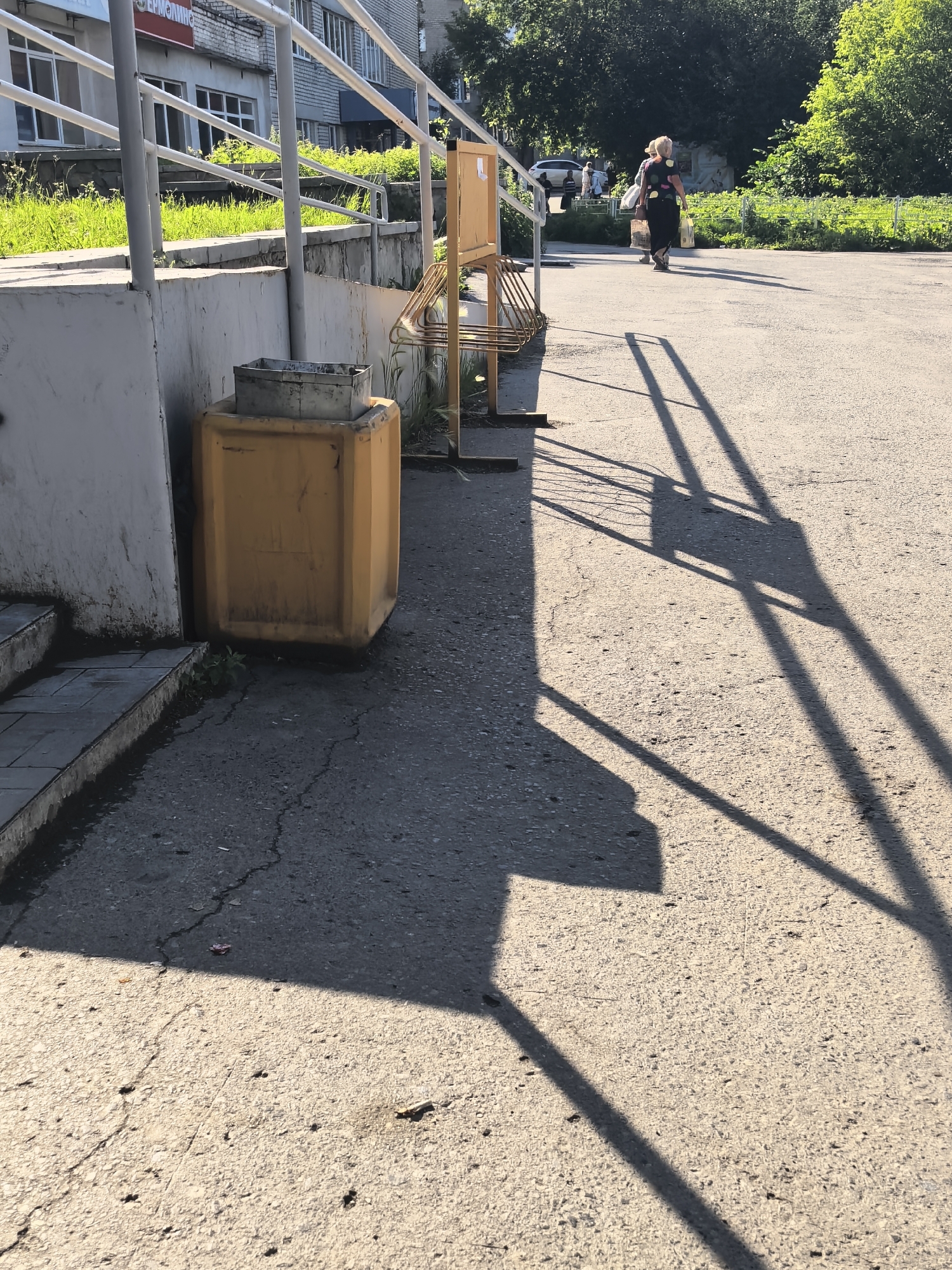 The image shows an outdoor scene with a woman walking away from the camera in the distance. She is carrying shopping bags and wearing a dark dress with colorful polka dots. The scene appears to be a sunny day, with long shadows cast by a railing and a bike rack. The ground is paved with asphalt, showing some cracks.
In the foreground, to the left, is a gray metal trash can on top of a large yellow plastic bin. Next to the bins are some concrete steps with grass growing between the tiles. A white concrete wall is also visible. Behind the wall and railing, there is a patch of vibrant green grass and some trees.
In the background, a light-colored car is parked on the side of the road, and another person is walking. A building with windows and a white facade is visible behind the greenery and railing. A sign with some text is partially visible on the building, but it is not clearly legible. The overall impression is a casual daytime scene in an urban or suburban environment.