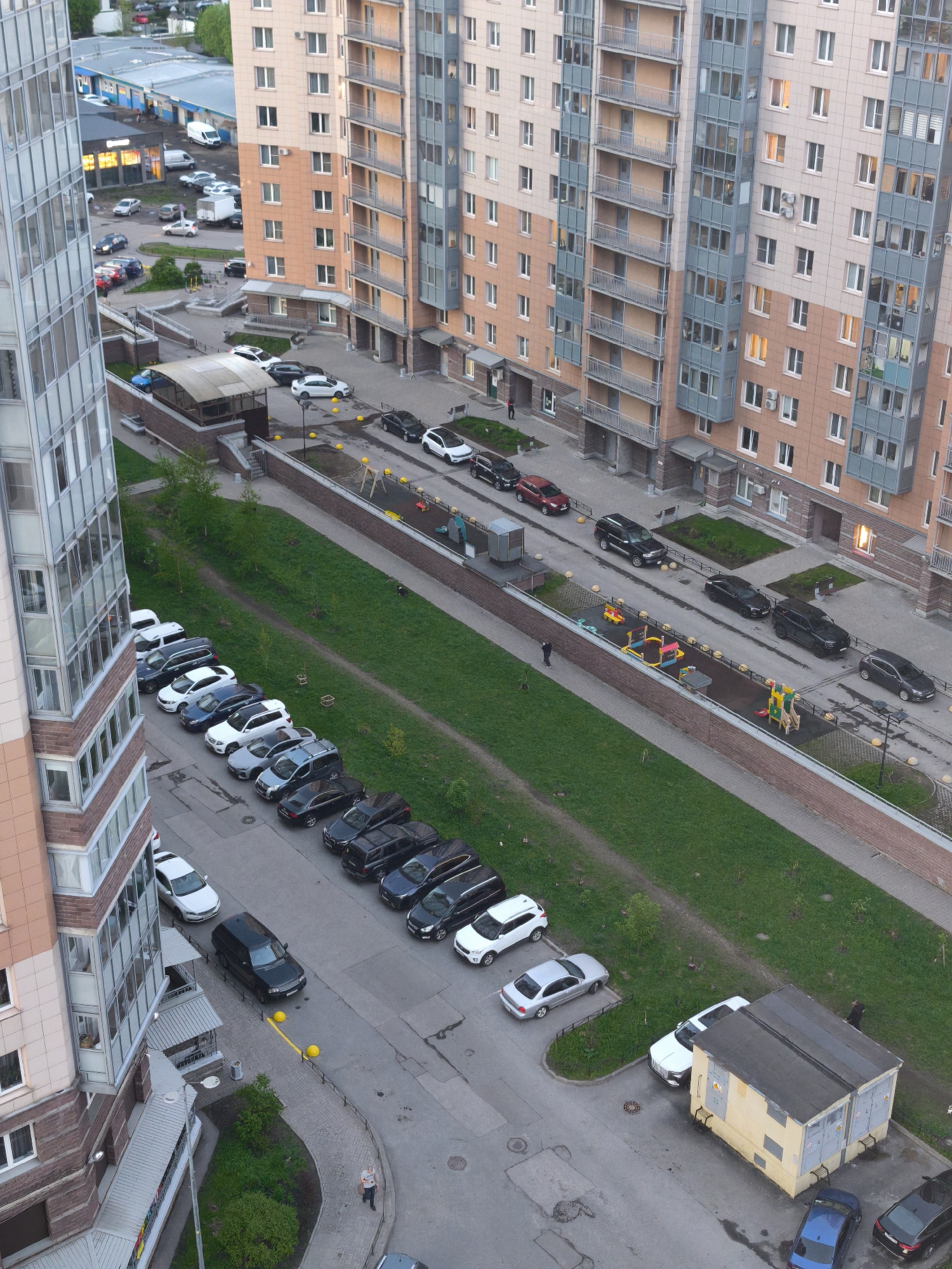 The image shows an aerial view of a residential area in Saint Petersburg, Russia. The scene is dominated by several tall apartment buildings with beige and blue facades, interspersed with balconies. Between the buildings, there are parking lots filled with various cars, including SUVs, sedans, and hatchbacks, predominantly in colors like white, black, and silver. Green grassy areas run through the complex, separating the parking areas and walkways. A children's playground with colorful equipment is visible alongside one of the buildings. A small yellow utility building sits near a parking area. Several people are scattered throughout the scene, mostly walking along the walkways or near parked cars. The overall environment suggests a densely populated urban setting with a mix of residential buildings, parking facilities, green spaces, and recreational areas.