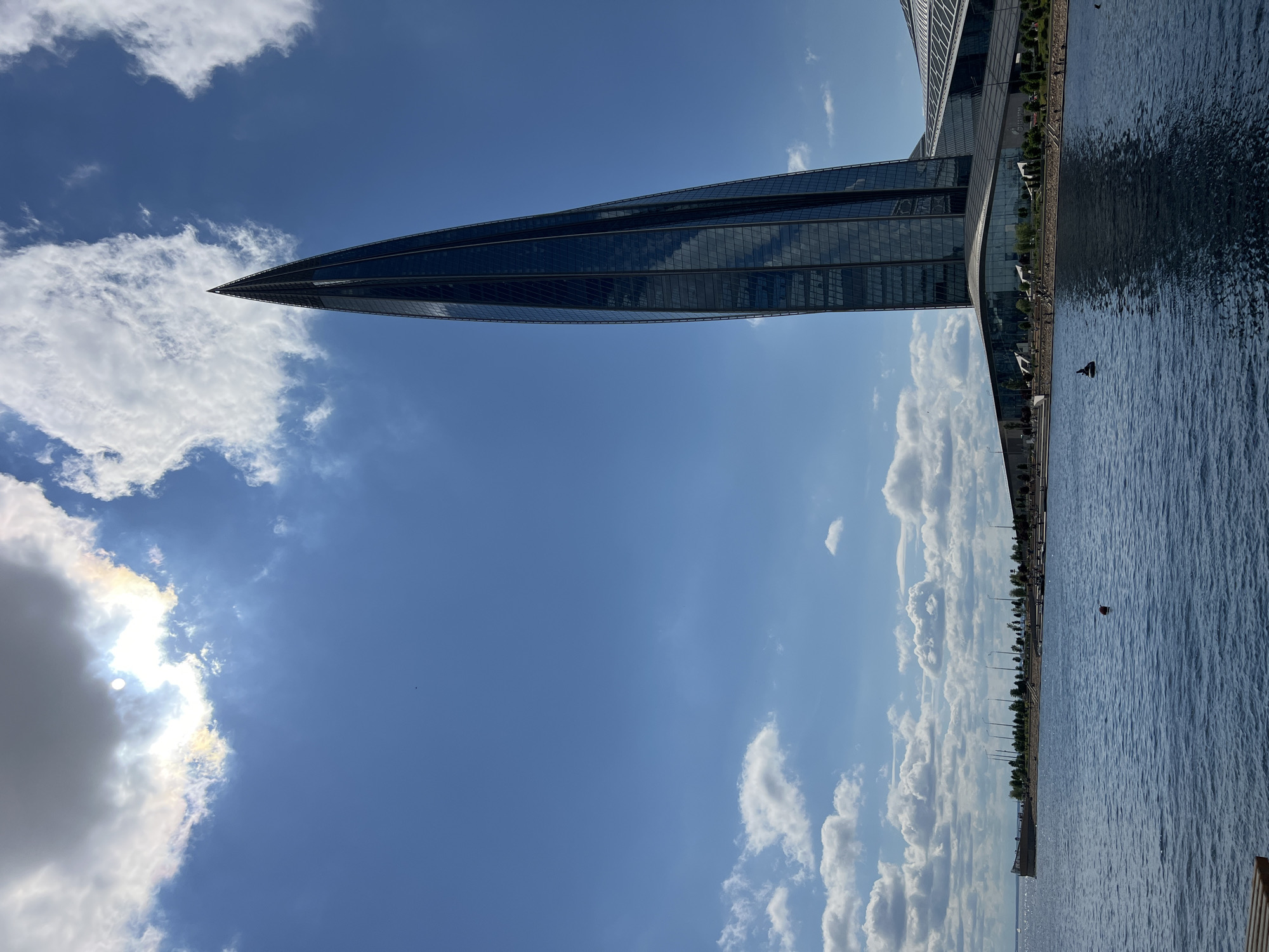 A tall, modern, dark glass skyscraper with a pointed top dominates the right side of the frame, leaning slightly to the left. The skyscraper's reflective surface mirrors the bright blue sky and scattered white clouds. In the foreground, a body of calm, dark blue water stretches across the bottom of the image. The water shows ripples and reflections of the sky and the building. Along the left bank, a shoreline is visible with a promenade, trees, and lampposts. Several people are scattered along the shoreline and in the water, some appearing to be sunbathing or walking. The sun is partially obscured by a large, gray cloud on the upper left, casting a bright glow. The scene depicts a sunny day in an urban waterfront area.