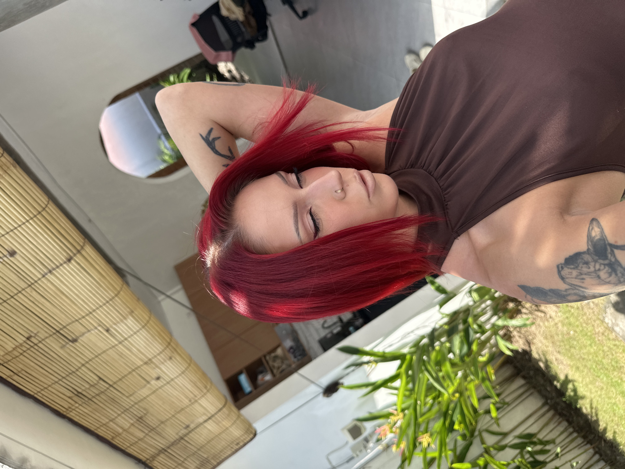 A woman with vibrant red hair lies supine, eyes closed, wearing a brown halter-style top. A small gold hoop adorns her nostril. Tattoos are visible on her arms, one a stylized antler design, the other an indistinct, possibly animalistic image. She is outdoors; in the background, greenery is seen near what appears to be a white wall and green grass. A bamboo awning-like structure is also visible. It can be determined, per the metadata, that the location is in North Kuta, Indonesia.
