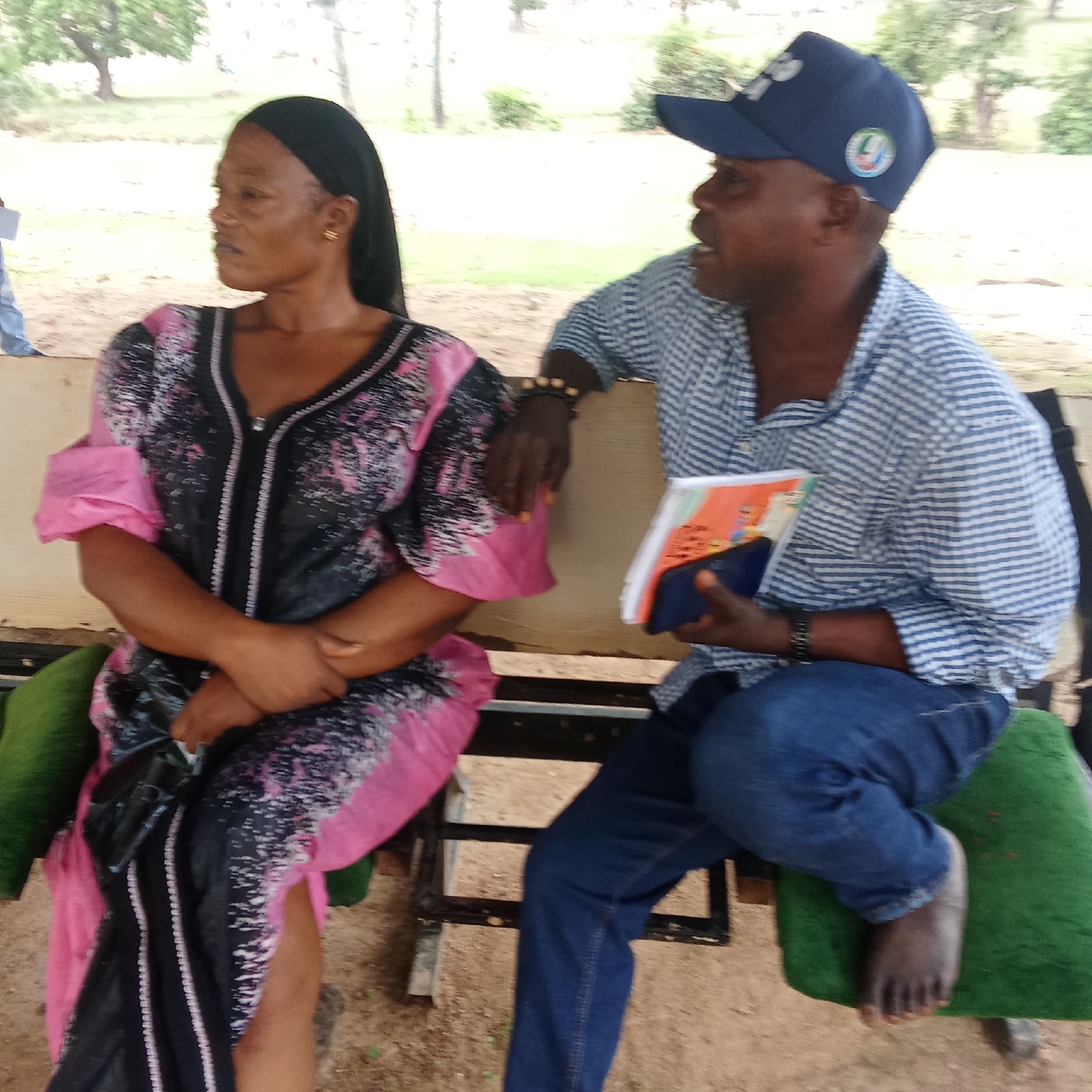 Two individuals are seated on a bench in an outdoor setting. A woman wearing a black and pink dress with her hair wrapped sits to the left, while a man in a blue and white striped shirt, blue jeans, and a dark blue baseball cap sits to the right. The man is holding a book and a dark object (possibly a phone or wallet). He is barefoot and appears to be speaking. The location is likely in Akwanga, Nigeria, based on the query. The bench has green cushions. The background includes trees and greenery, suggesting a park or outdoor recreational area.