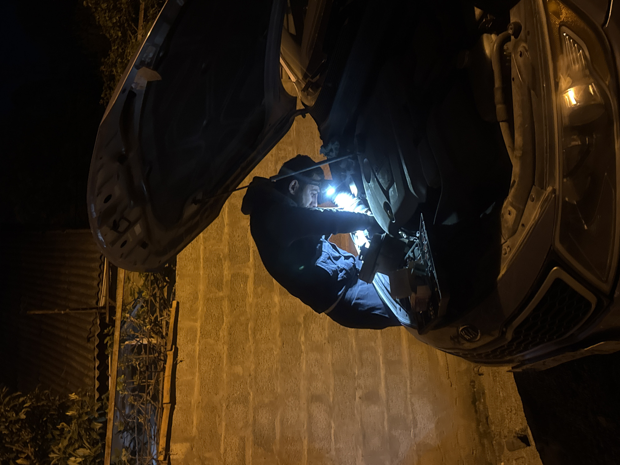 A man repairs a car at night near a building in Tbilisi, Georgia. The man is wearing dark clothing and a headlamp. The car's hood is open, and the man appears to be working on the engine. A wall is visible behind the man, and there is a section of corrugated metal and foliage to the left of the man.
