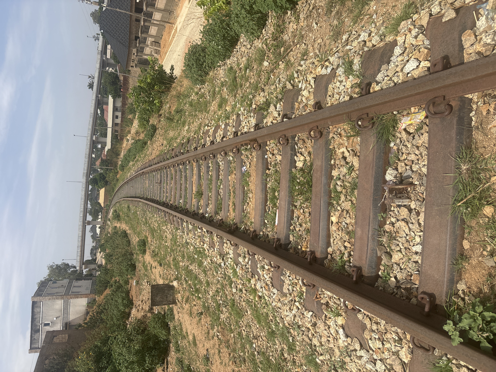 The image depicts a section of railway tracks curving into the distance. The tracks are rusty and show signs of disuse. The railway bed is composed of gravel and sparse vegetation. Buildings and a bridge-like structure are visible in the background, indicating an urban environment. Based on the prompt, the location is identified as Jos, Nigeria. No people are visible in the image.