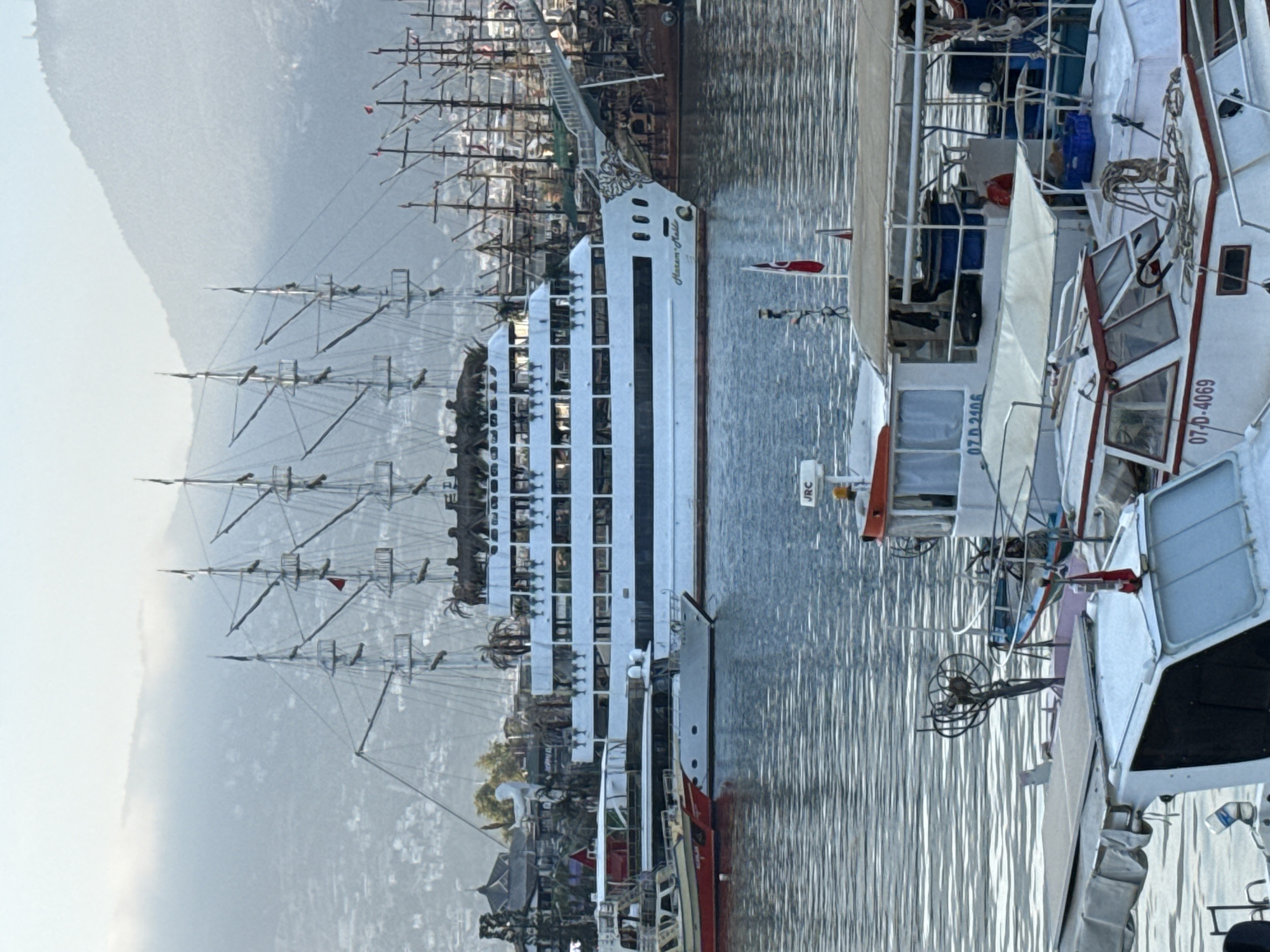 The media file depicts a harbor scene in Alanya, Turkey, with several vessels docked. Dominating the foreground is a large, modern white ship with multiple decks and rows of windows. To its right, part of another white boat with red trim is visible, featuring windows and a davit. Further to the right, another boat is partially seen, also with white and red elements. In the background, a classic sailing ship with multiple masts and sails is prominent, along with other sailboats and masts. The water is calm and reflects the sky and the boats. The scene appears to be set during daylight, likely morning or afternoon, given the lighting. The overall atmosphere is serene.
Visible text includes "MARINA MALTA" on the side of the large white ship, and registration numbers "07-D-2106" and "07-D-4069" on two of the boats. There is also a label "JRC" on one of the smaller vessels.