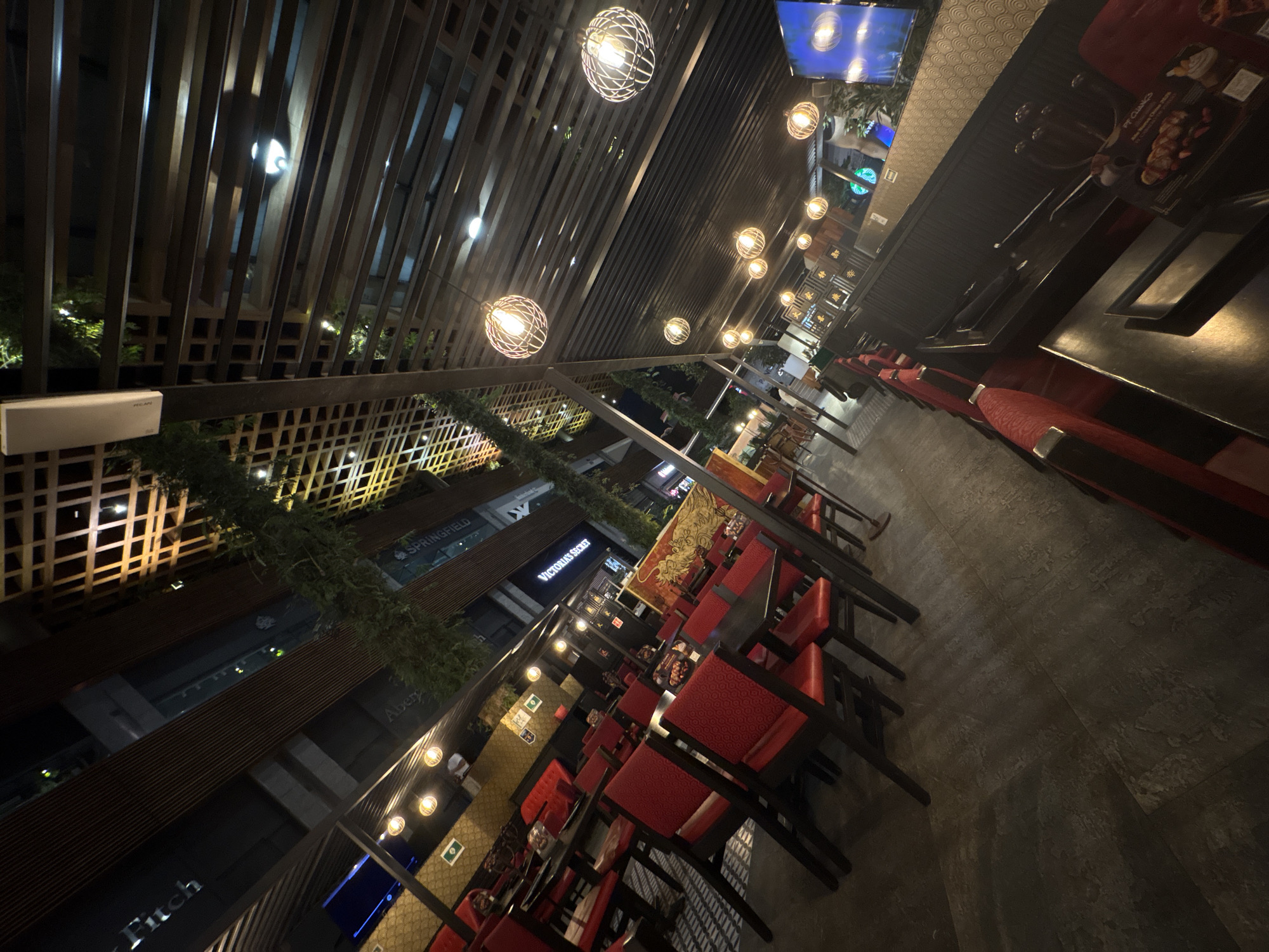 The scene depicts an outdoor dining area of a restaurant, identified as P.F. Chang's by a visible menu, located within the Angelópolis Lifestyle Center in Puebla, Mexico, at night. Overhead, a dark, slatted wooden pergola structure is adorned with artificial green foliage and numerous spherical, cage-style pendant lights featuring exposed filament bulbs. Recessed white lights and a white rectangular communication device are also visible on the ceiling structure.
The restaurant features dark, square-top tables paired with dark-framed chairs upholstered in red fabric. Some tables are positioned alongside banquette-style seating, also in red. The flooring is dark gray, textured tile. A wall section to the left displays a gold geometric pattern with small green, white, and red flags, likely decorative. Another prominent wall feature, visible to the right, showcases a large golden dragon motif over a red and gold patterned background, along with Chinese characters.
In the background, illuminated storefronts of retail businesses such as "Victoria's Secret," "Springfield," and partially "Abercrombie & Fitch" are visible, confirming a commercial shopping center environment. A single individual is discernible in the mid-ground, standing or walking near the rear of the dining area. The restaurant appears sparsely populated with patrons. The overall lighting scheme is warm, primarily from the suspended fixtures, contrasted with the brighter white lights of the distant storefronts.