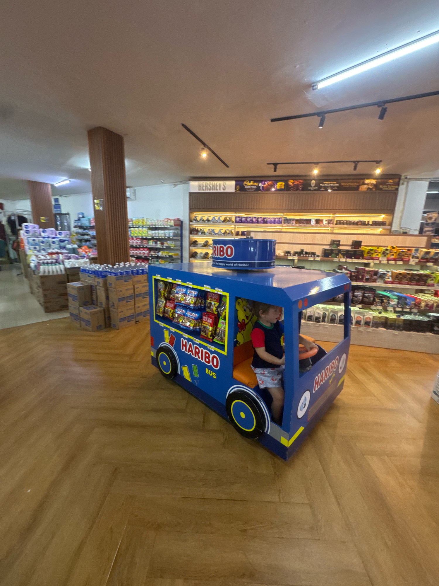 A young Caucasian child, wearing a blue and red shirt and patterned shorts, sits inside a blue Haribo bus-shaped ride-on toy in a store aisle. The child is facing forward, with their hands on the steering wheel. The toy bus has "HARIBO BUS" printed on its side in white lettering and features a yellow cartoon bear image. Various shelves stocked with products, including bottled water and packaged goods, are visible in the background. A wooden herringbone floor covers the ground. The scene appears to be a retail environment, possibly a convenience store or supermarket, located in Kuta Selatan, Indonesia.