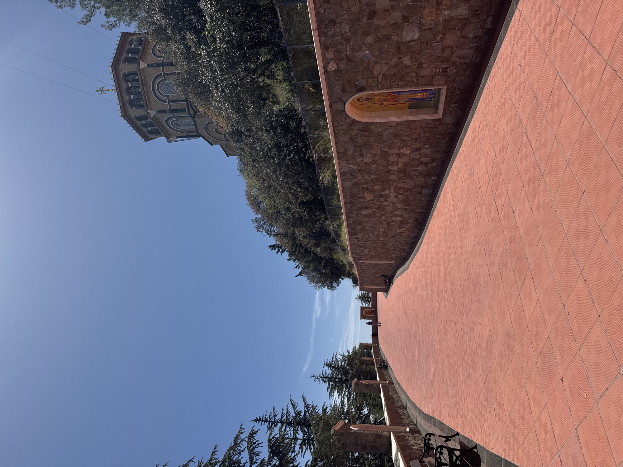 The image shows a tiled pedestrian walkway in Tsakistra, Cyprus. A stone wall runs along one side of the walkway, featuring a mosaic depicting a religious figure. On the other side are benches and trees. In the distance, people can be seen walking along the path. A building with a cross on top is visible through the trees on the left. The sky is clear and blue.