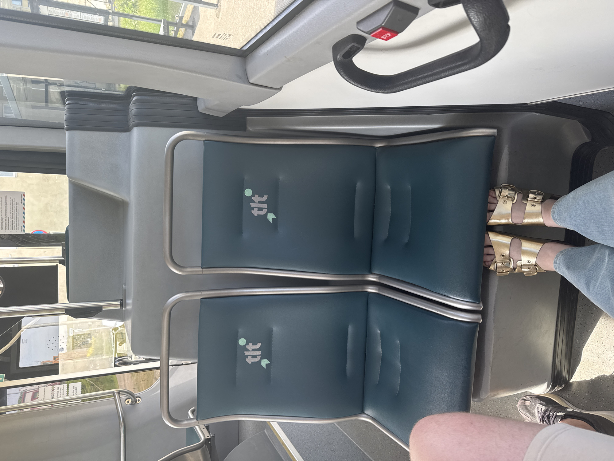 The image shows the interior of a public transportation vehicle, likely a bus or tram, in Tallinn, Estonia. Two empty teal-colored seats are prominently featured in the foreground, each with a "tlt" logo, which stands for Tallinna Linnatransport, the public transport operator of Tallinn. A person's legs and feet are visible in the lower portion of the image. The person on the left is wearing grey sneakers and light-colored shorts, while the person on the right is wearing gold sandals and blue jeans. The seats are positioned behind a grey console. The environment appears to be a sunny day, with sunlight filtering through the windows. There is a black handrail with a red "STOP" button visible to the right of the seats. The overall scene depicts a moment of pause or travel within the city's public transit system.