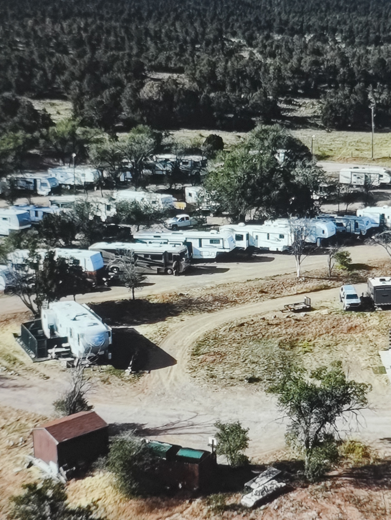 The image depicts an aerial view of an outdoor area with numerous recreational vehicles, including travel trailers and motorhomes, parked in rows. The vehicles are of various sizes and colors, with some appearing to be newer models and others older. The ground is a mix of dirt roads and gravel, with patches of dry grass and scattered trees. In the foreground, there is a small, dark red shed with a brown roof. Further into the scene, there are more RVs, some with awnings extended, and a few cars. The background consists of a dense forest with a hilly terrain. There are no visible people in the image. The scene appears to be a campground or RV park. The request for a location in Yarmolyntsi, Ukraine is not supported by any visual evidence in the image.