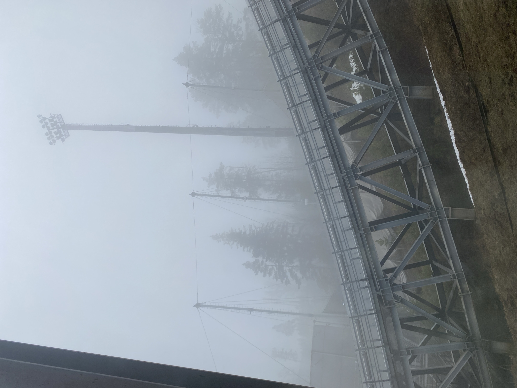 The image displays an outdoor scene characterized by heavy fog and large man-made structures. A prominent feature on the right is a substantial dark grey metal truss structure, composed of intricate beams and girders, extending horizontally across the frame. This industrial-looking framework suggests it could be part of a large construction, such as a bridge, a stadium support, or an industrial facility. To its immediate right, a flat, brownish area resembling dry grass or earth is visible.
In the midground and extending towards the left, several tall, slender poles or towers rise vertically, partially disappearing into the dense fog. One tower on the far left is clearly topped with a cluster of bright floodlights, indicative of a lighting mast for a sports arena or outdoor venue. Other similar poles are faintly visible behind it, their details softened and obscured by the atmospheric conditions.
The background is almost completely enveloped in thick, white fog, through which only the faint, dark silhouettes of evergreen trees can be discerned, hinting at a natural landscape beyond the structures. The pervasive mist creates a muted, monochromatic palette and diffuses the light, making it difficult to ascertain the precise time of day, though it appears to be daylight.
There is no visible human activity or text in the image. The scene conveys a sense of stillness, isolation, and mystery due to the overwhelming fog, set against what appears to be an industrial or sports complex environment in the Прикамский region of Russia.