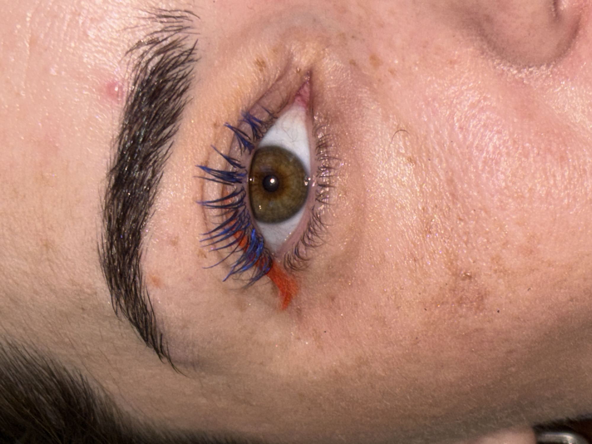 A close-up, eye-level, medium shot shows the right eye of a person with fair skin, freckles, and dark eyebrows. The eye is open and looking slightly to the left of the camera. The iris is a hazel-green color. The person is wearing blue and orange mascara on their upper lashes and a subtle orange eyeliner on the inner corner of the eye. There is a small red blemish on the forehead above the eyebrow. The skin has a slight shimmer, possibly from highlighter or glitter. The background is out of focus and shows hints of what appears to be an indoor setting. No details about the location in Sinyavino, Russia are present in the image.