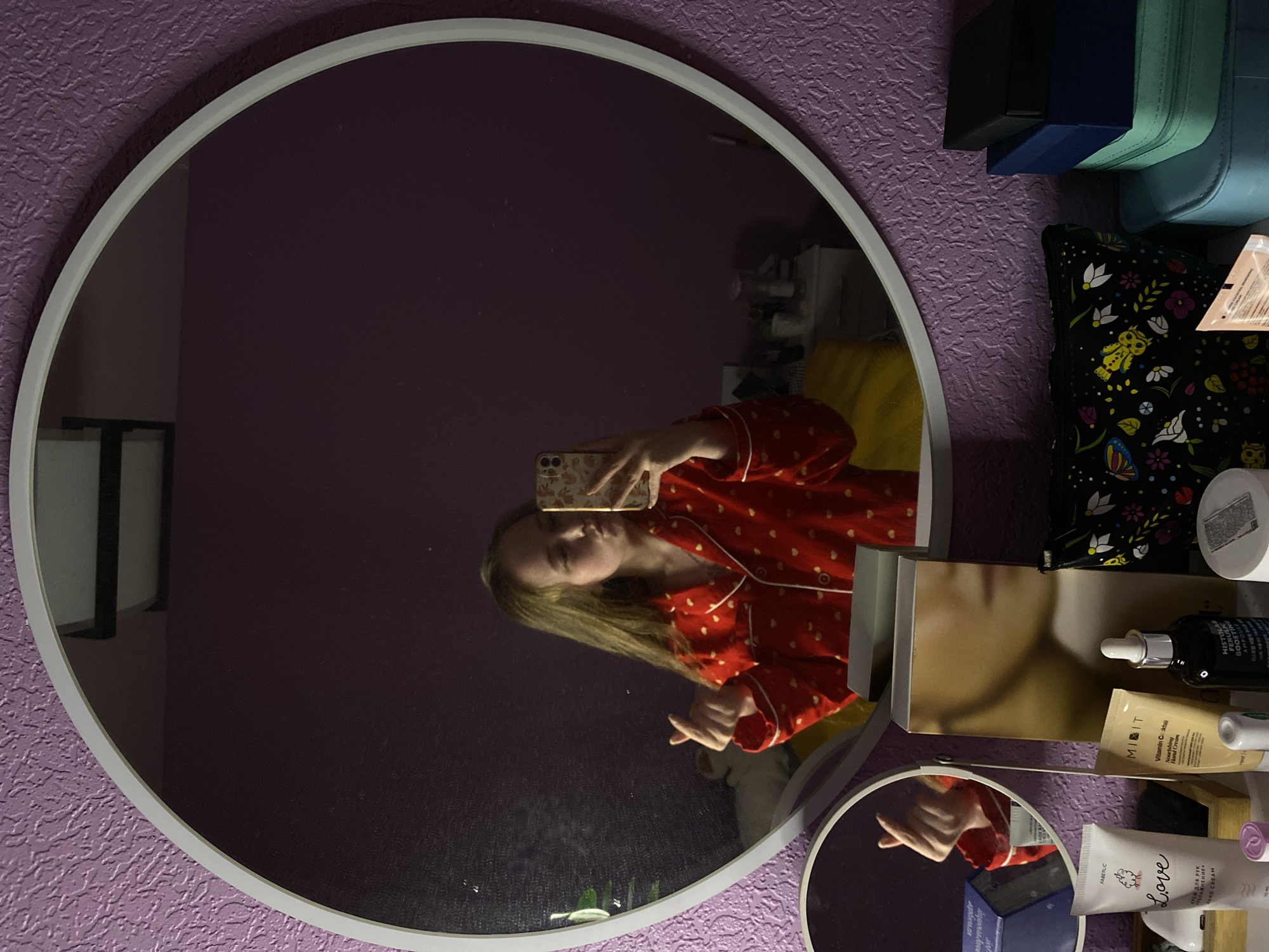 A reflection in a round mirror shows a woman holding a phone, presumably taking a selfie. She is wearing red pajamas with white hearts. The mirror hangs on a textured purple wall. Beside the mirror, on the right, are various cosmetic products, including tubes and bottles of creams and serums. Some containers bear the labels "FABERLIC Love", "MIDIT Vitamin Cocktail", and "HISTOLAB FERULIC BOOSTING AMPOULE". A black bag with a floral pattern sits among the cosmetics. The reflection in the mirror shows a bed with yellow sheets. There is also a smaller round mirror below the main one, reflecting the woman's arm and sleeve. The location is likely in Russia, Saint Petersburg due to the name of the cosmetic brands being in Russian.
