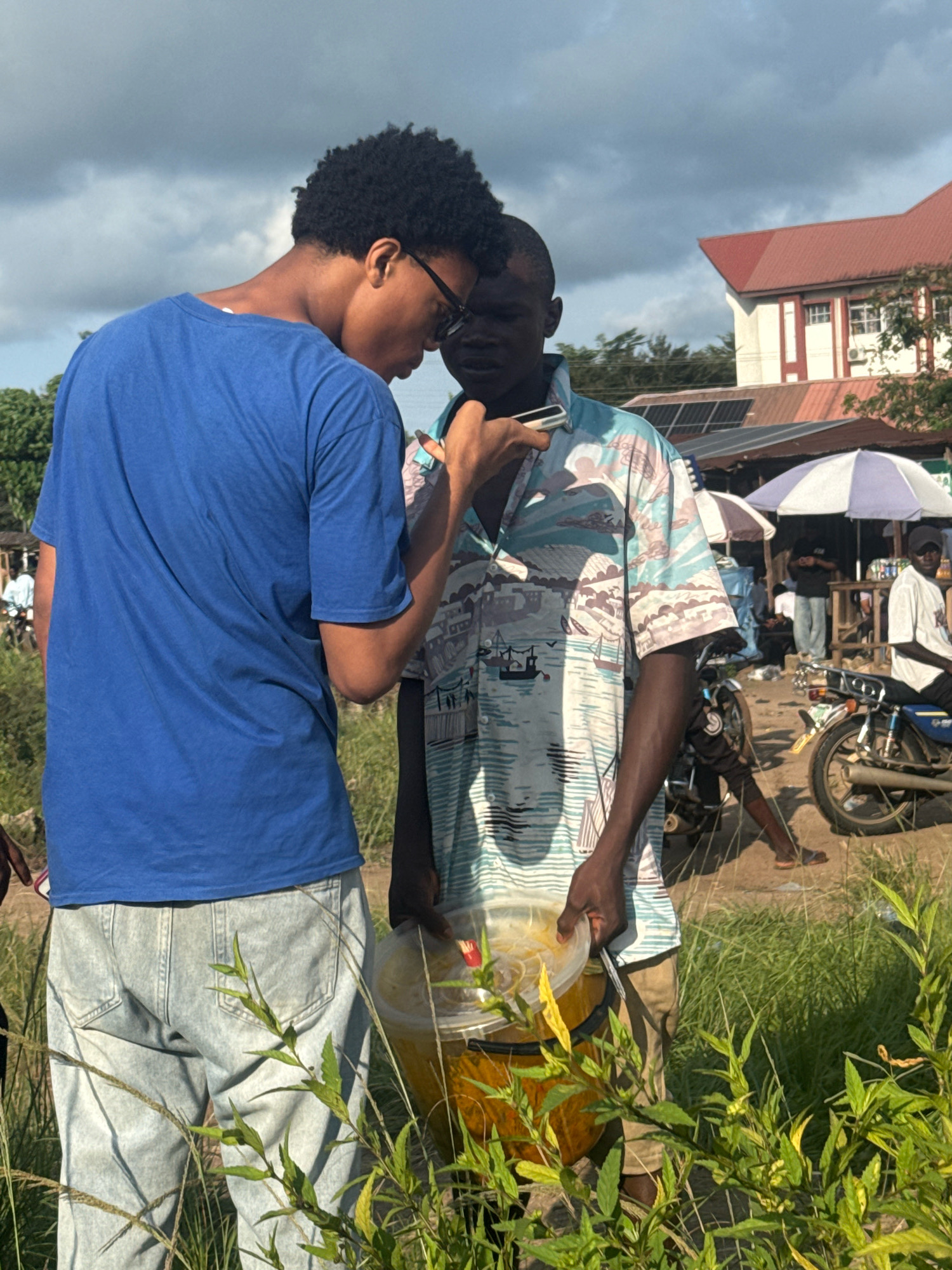 In Ihiagwa, Nigeria, two young men are engaged in conversation under a cloudy sky. The man on the left, wearing a blue t-shirt and light-wash jeans, is looking at his phone. The man on the right, in a patterned short-sleeved shirt and khaki pants, holds a clear plastic bucket filled with a yellow liquid. Behind them, a building with a red roof is visible, along with market stalls, umbrellas, and parked motorcycles. The scene is set in an outdoor environment with tall green grass in the foreground.