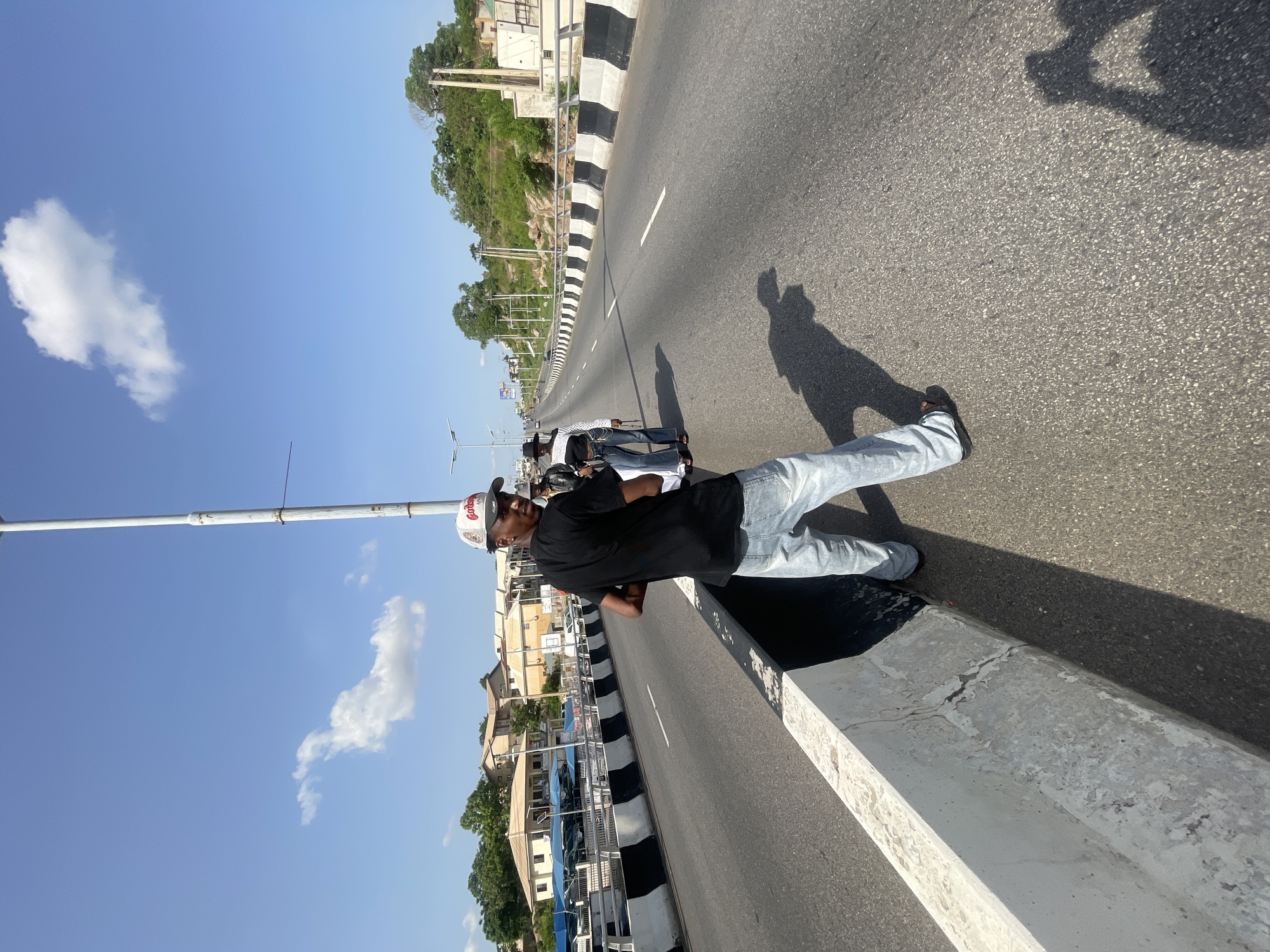 A man in a black t-shirt, light blue jeans, and a baseball cap stands on the edge of a highway overpass. He is on the right side of the road, leaning slightly over a white concrete barrier. The road is paved with asphalt and marked with white lines. A black and white striped barrier runs along the side of the road. In the background, there are buildings and trees. The sky is blue with some clouds. The man's shadow is cast on the road. The image was taken on a sunny day in Lokoja, Nigeria.