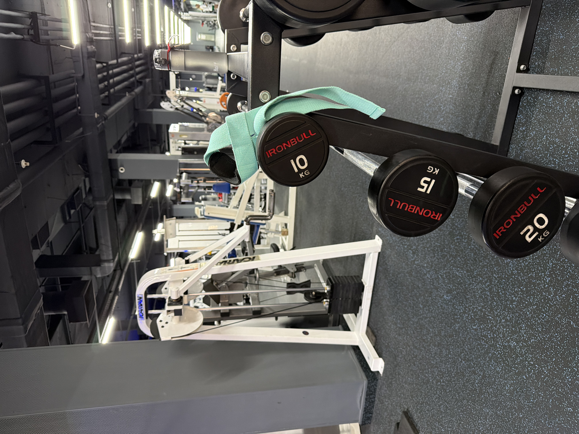 The image shows a weight rack with dumbbells ranging from 10 kg to 20 kg, manufactured by IronBull, with a light green wrist strap draped over the 10 kg dumbbell. The rack is in a fitness center, as suggested by the exercise machines visible in the background. The floor is a textured, speckled gray, and the gym has high ceilings with exposed pipes and lighting fixtures. The location is inferred to be in Kazan, Russia due to the branding on the equipment.