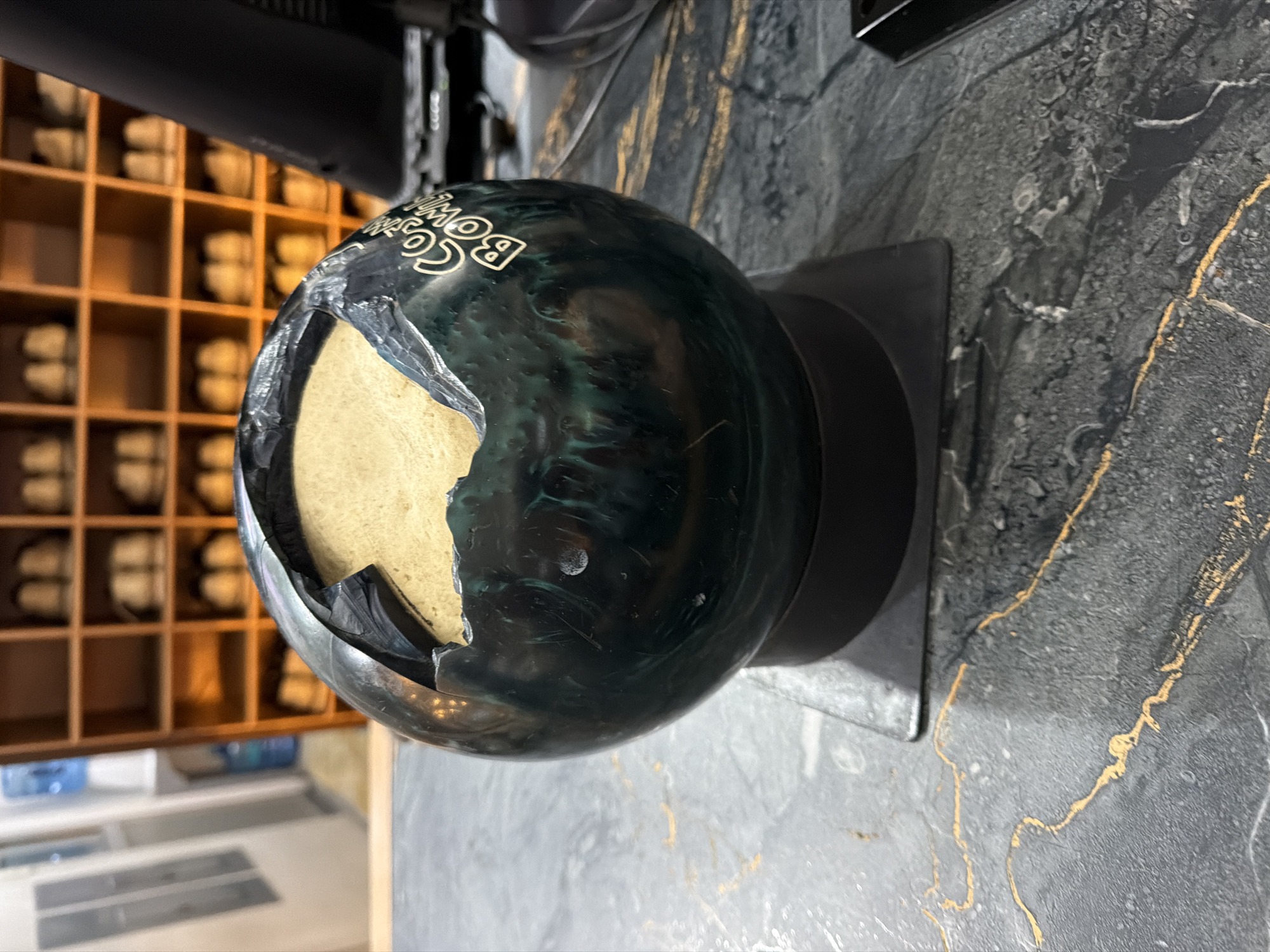 A severely damaged bowling ball, predominantly dark teal with black swirling patterns, is centrally displayed on a black square stand. A large portion of the ball's upper shell is fractured and missing, exposing a light tan, granular inner core. White text, "Cosmic Bowl," is visible on the intact side of the ball. The stand rests on a dark gray countertop with distinct golden-orange veining. To the right, a dark computer monitor and a segment of a keyboard are partially visible. The background features a large, light brown wooden rack with multiple square compartments, each containing light-colored bowling shoes. A blurred white doorway and a blue water cooler are discernible in the far-left background. The scene depicts a reception or shoe rental area within a bowling alley in Puebla, Mexico.