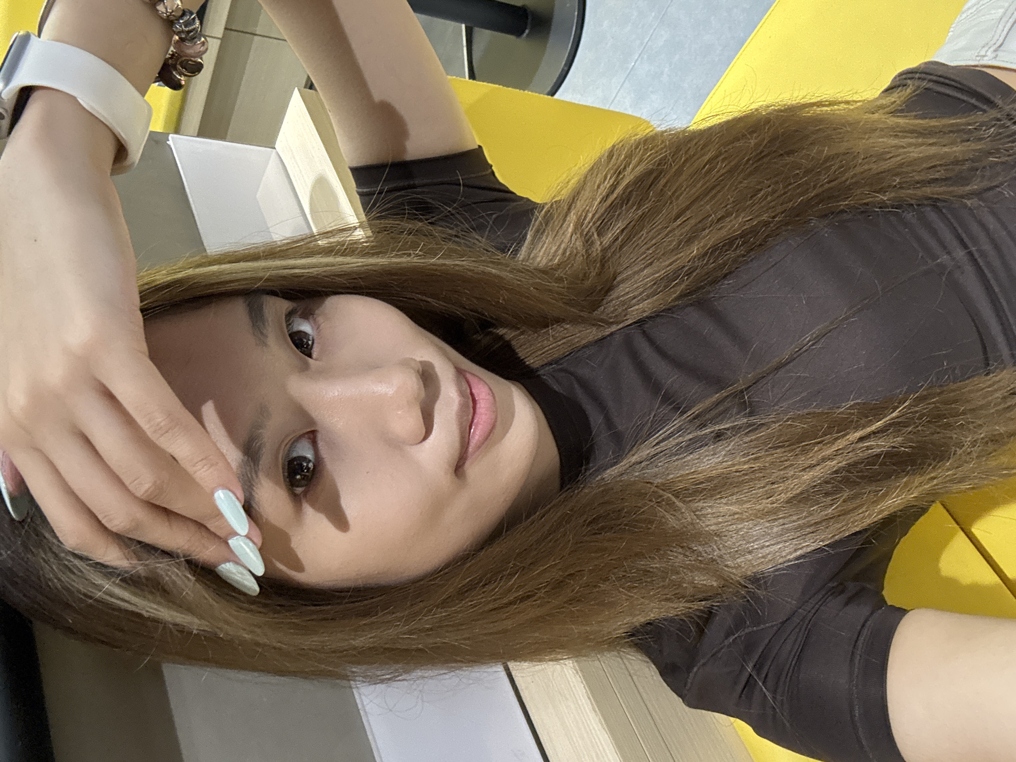 A woman with light brown hair is lying on a yellow surface, possibly a seat or cushion. She is wearing a dark brown, fitted top with short sleeves. Her left hand is raised towards her forehead, displaying light blue-green nails. A white smartwatch is visible on her left wrist, along with a multi-colored bracelet. The background includes elements of an interior space with gray and white surfaces. Given the context request, the general location would be in the Yuzhong District of China, but no specific street address can be derived from this single image.