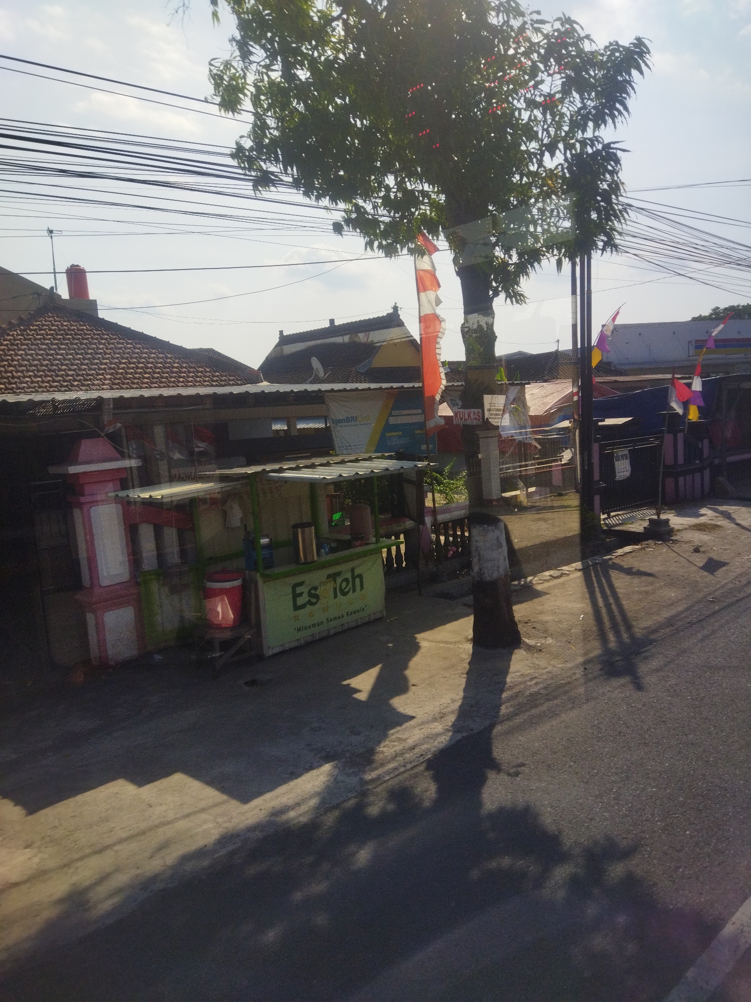 This image depicts a street scene in Nambangan, Indonesia, under a clear sky with the sun casting long shadows. In the foreground, a paved road is visible with a concrete sidewalk or paved area to its left, partially covered by a patterned shadow. A small food stall with a green banner and the words "Es Teh" (Iced Tea) is set up on this paved area. Next to the food stall is a decorative pink and white structure with multiple levels, possibly a display or a planter. Behind the stall, a tree with green foliage and some red fruit or decorations stands tall, its trunk wrapped with white and red striped fabric, resembling a flag. Further back, various buildings with tiled roofs are visible, along with utility poles and a dense network of overhead electrical wires. Flags, including Indonesian flags and other colorful banners, are attached to poles and buildings. The overall scene suggests a commercial or residential area in a tropical climate, with elements of everyday life such as street vending and visible infrastructure.