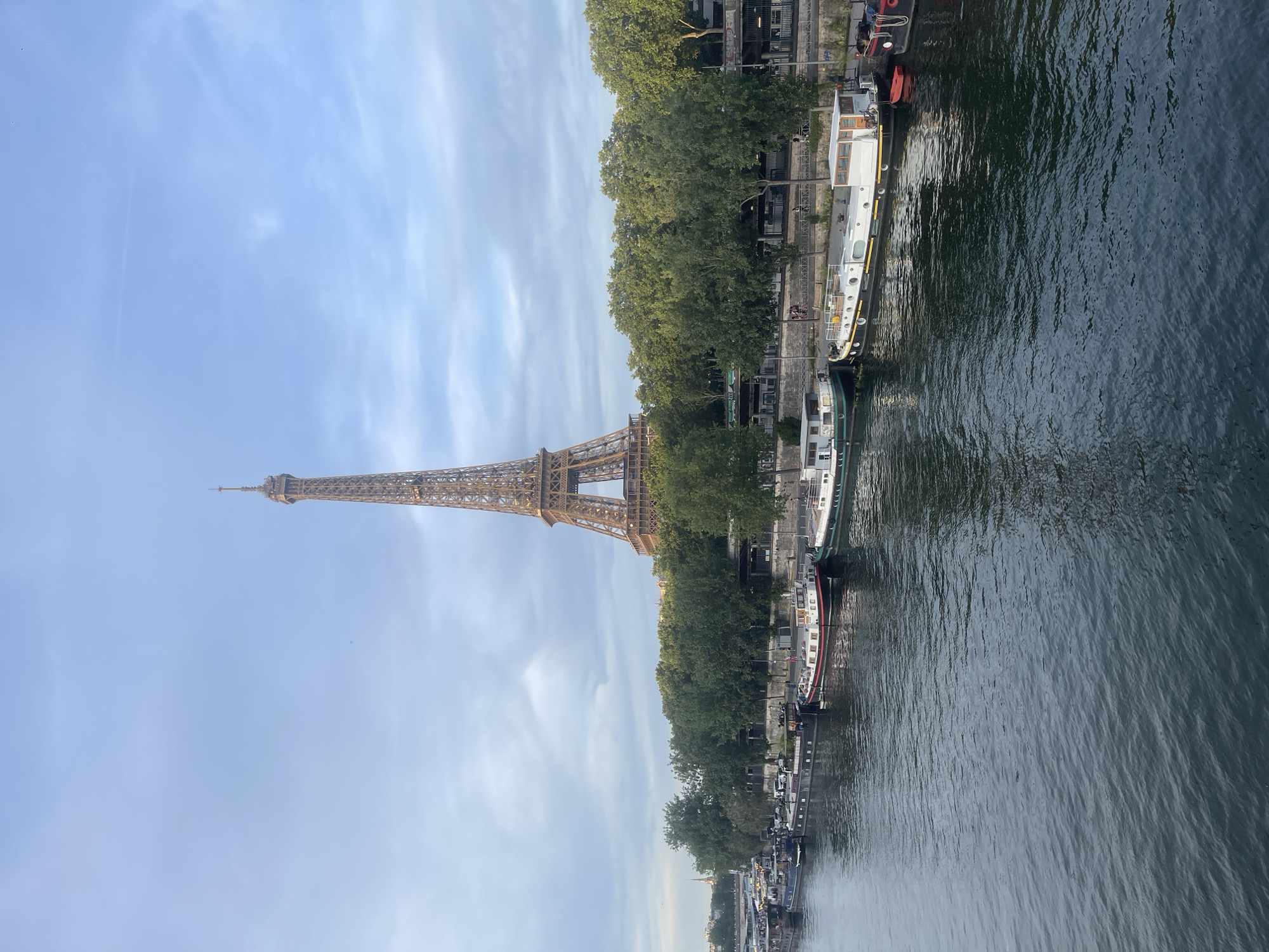 This image captures a serene view of the Eiffel Tower in Paris, France, as seen from the Seine River. The iconic iron lattice tower dominates the upper portion of the frame, bathed in the soft light of a partly cloudy sky. In the foreground, the tranquil waters of the Seine reflect the muted hues of the sky and surrounding trees.
Along the riverbank, a line of houseboats, or barges, are moored, adding a touch of residential life to the urban landscape. These boats vary in size and design, with some featuring white hulls and others darker tones, some with red accents. They are neatly arranged, creating a picturesque scene against the backdrop of lush green trees that line the embankment.
Beyond the trees, glimpses of Parisian architecture can be seen, hinting at the vibrant city surrounding this peaceful waterway. The overall atmosphere is one of calm and beauty, with the timeless Eiffel Tower standing as a silent witness to the flow of life along the river. There are no visible people in this particular frame, focusing the viewer's attention on the architectural marvel and the natural elements of the scene.