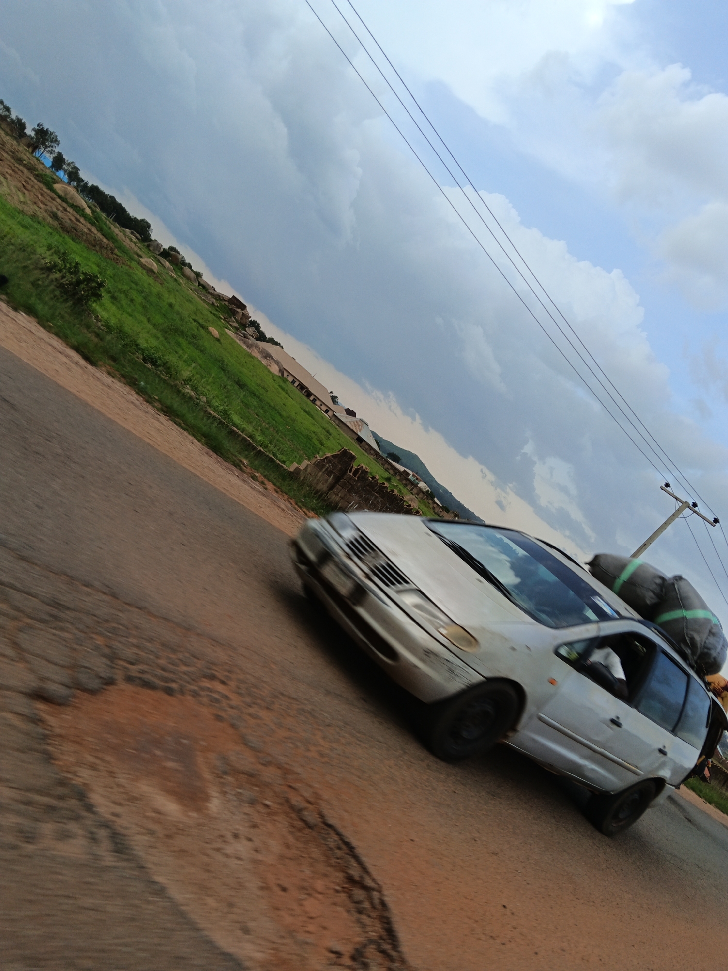 A silver minivan, laden with luggage secured to its roof, travels along a cracked and weathered asphalt road. The car is angled in motion, with the front on the left side of the frame and the back extending towards the right. The luggage on the roof appears to be in large, dark bags, possibly for travel. The road surface shows significant wear, with patches of exposed dirt and asphalt missing.
To the left of the road, a grassy hillside rises, dotted with large rocks and sparse trees. In the distance, structures are visible, including a low, stone wall and buildings with tiled roofs. The sky above is a dramatic mix of dark, heavy clouds and patches of bright blue, suggesting an impending storm or recent rain. Power lines stretch across the upper portion of the frame, supported by a utility pole visible on the right side.
The overall impression is one of travel and perhaps a journey through a rural or developing area. The lighting suggests it could be late afternoon or early evening, with the dramatic cloud cover creating a sense of mood. No visible text is present in the image.