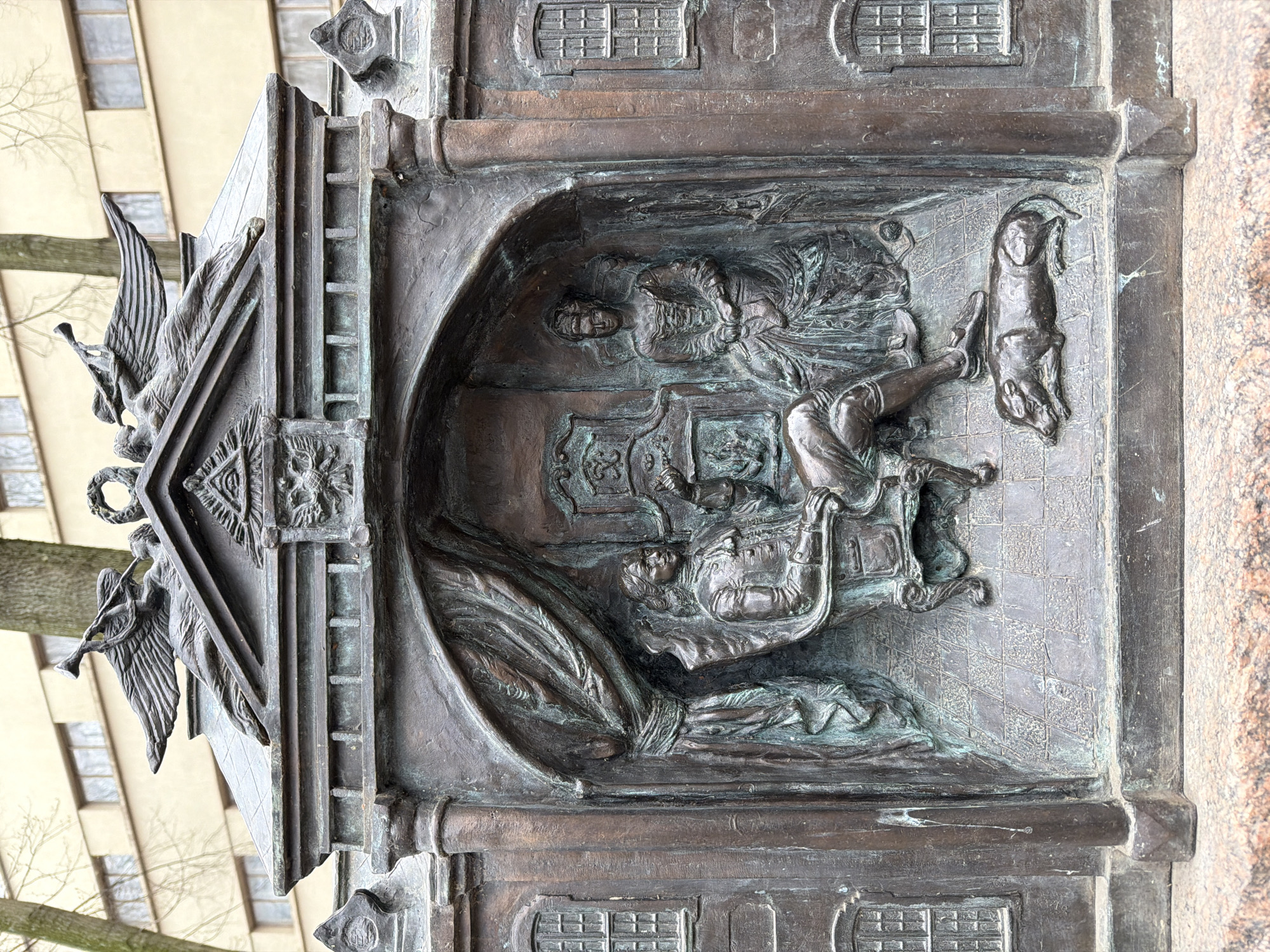 The image depicts a bronze relief carving from a monument. The relief is an interior scene. A man sits on a chair with his legs extended and his feet resting on the wall next to a hanging animal. A figure is lying on a draped surface at the bottom of the carving. Another figure is positioned above the seated man. The scene takes place in an enclosed room. The surrounding frame shows windows at the top and bottom. On the left of the framed relief is a carved heraldic design with winged figures and decorative elements.
Based on the style and iconography, this is likely a detail from one of the monuments in Saint Petersburg, Russia, where such commemorative bronze work is common.