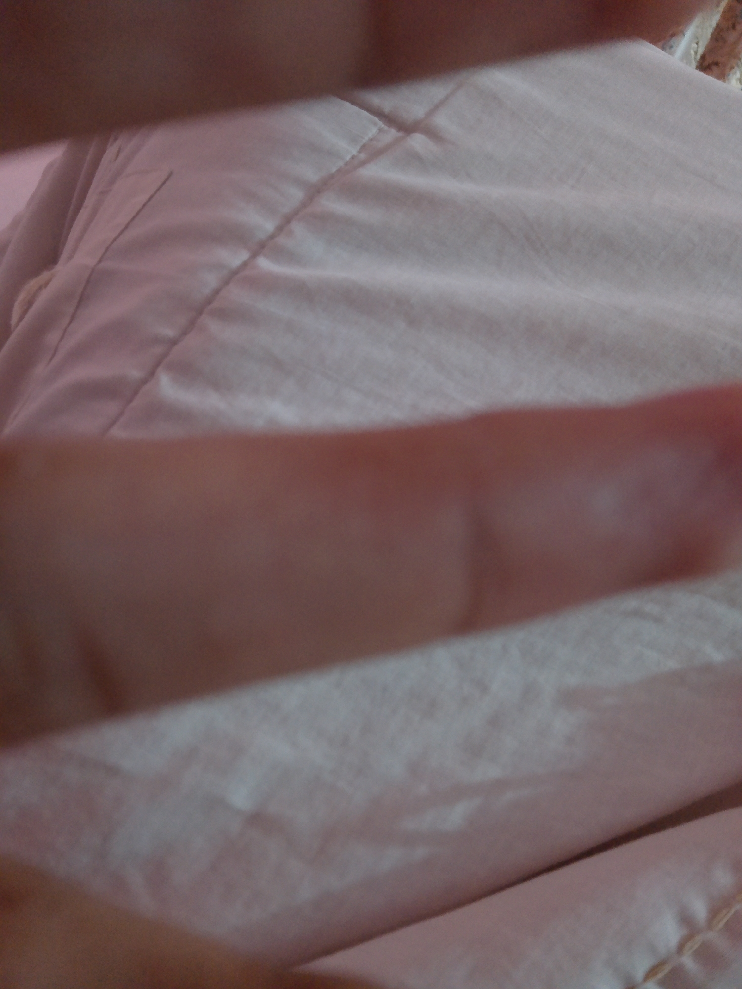 The image is a close-up, out-of-focus shot of a person's hand in front of a pink quilted fabric. The hand is large and fills the bottom half of the frame, with skin tones ranging from light to medium brown. The fabric is draped and creased, with visible stitching lines creating a pattern. The lighting appears soft, casting subtle shadows. The location in Mansehra, Pakistan, cannot be determined from the visual information provided.