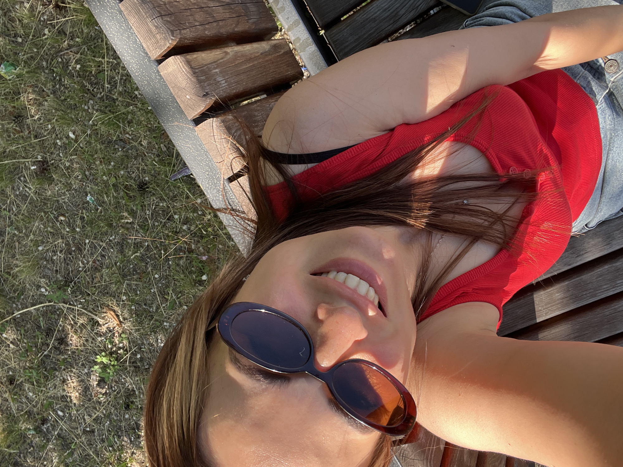 A woman with brown hair and sunglasses is taking a selfie on a wooden bench outdoors. She is smiling and wearing a red tank top and blue jeans. The background is a grassy area with trees.