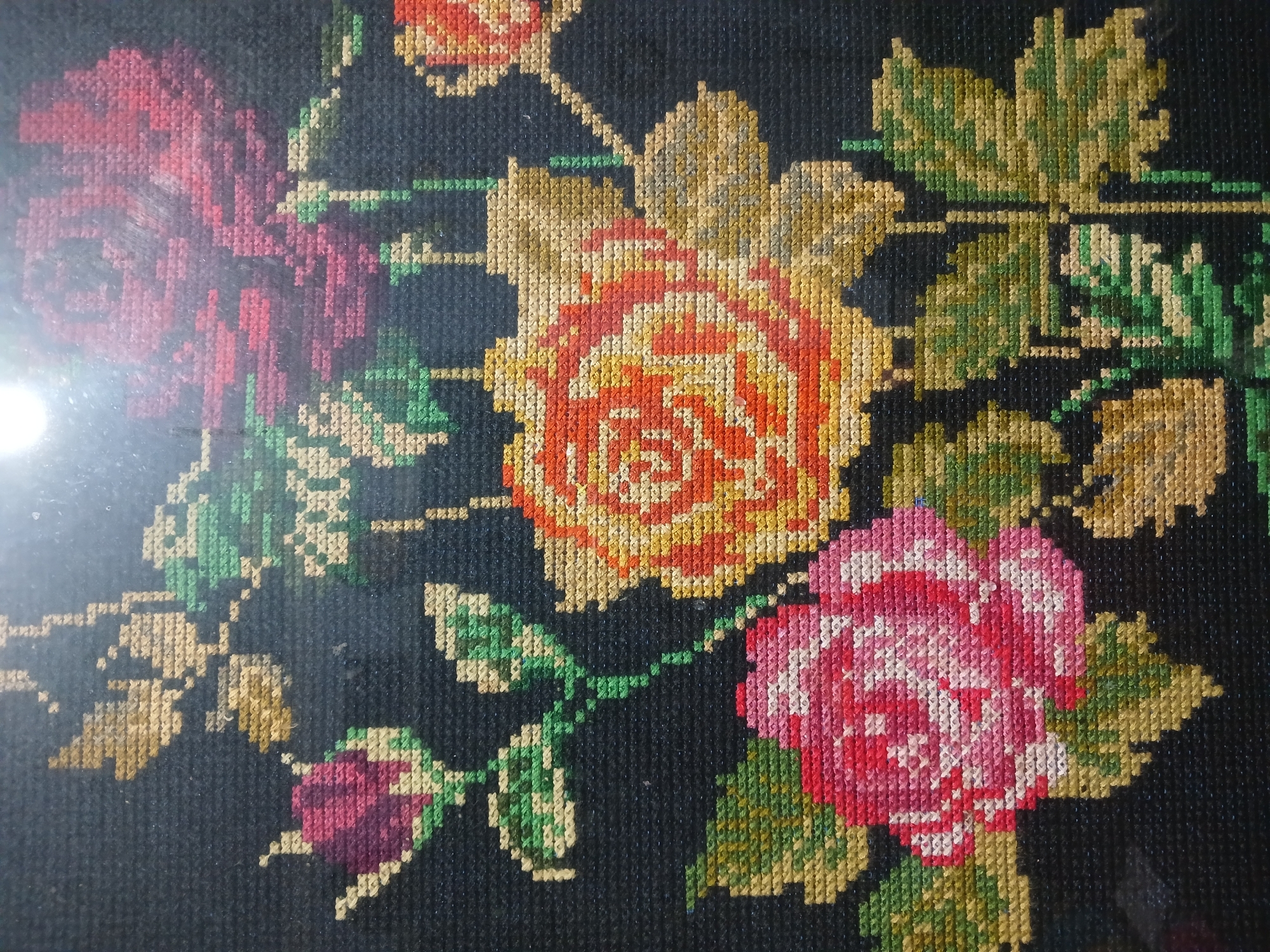 The image is a close-up photograph of a cross-stitch embroidery depicting a floral arrangement on a black background. The embroidery features several roses in various stages of bloom, rendered in shades of red, pink, orange, and yellow. Green and golden-brown threads are used to create the leaves and stems. The stitches are clearly visible, highlighting the texture of the textile art. The lighting in the image suggests it might be displayed under artificial light, as there is a bright reflection in the upper portion of the photograph. The image does not contain any information about its location in Randuagung, Indonesia, or any people, actions, or interactions. It is a static representation of a decorative handicraft.