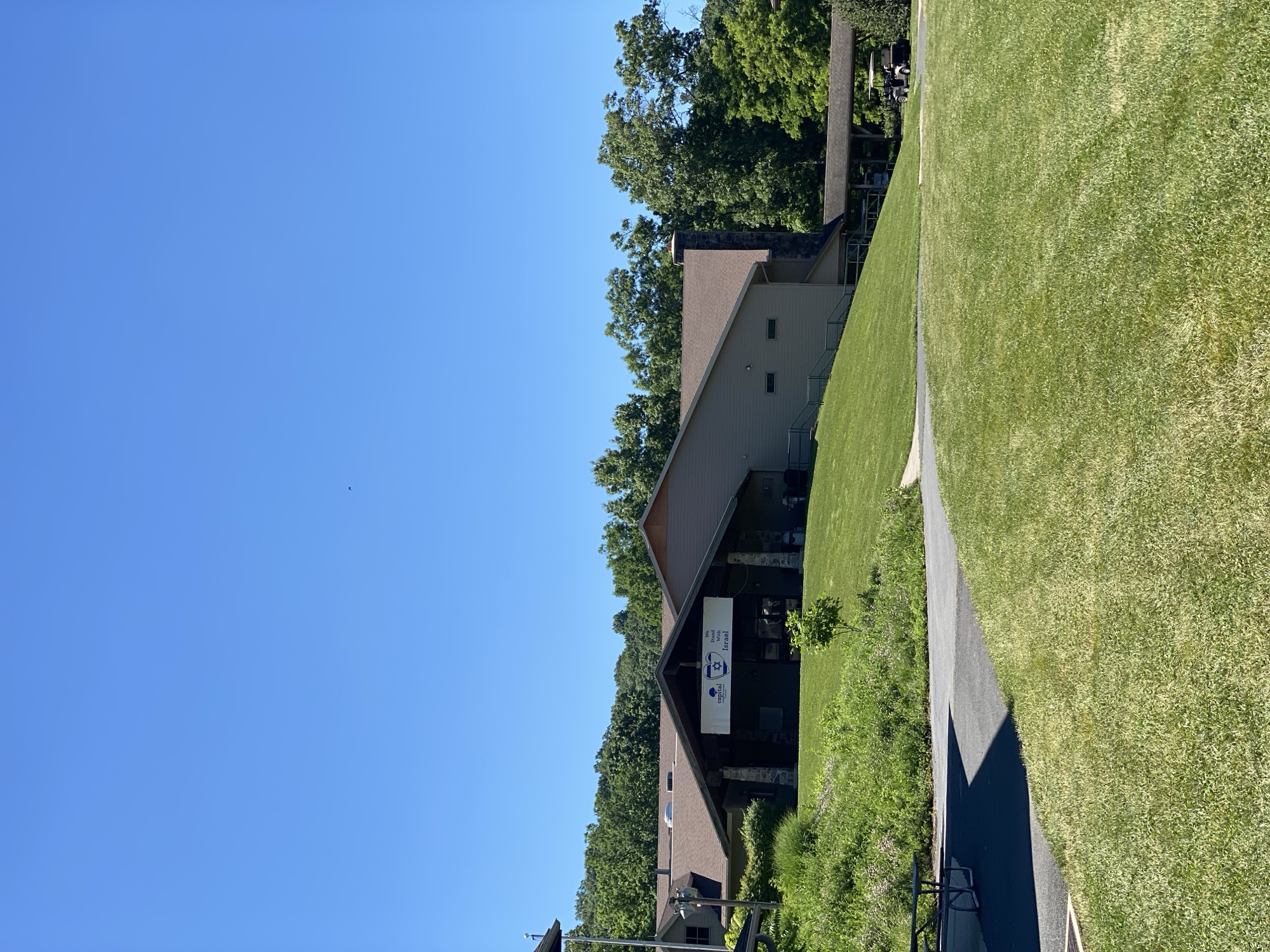 The image depicts a building with a brown roof and tan siding, situated on a grassy hill under a clear blue sky. A banner hangs over the entrance of the building, featuring the words "We Stand With Israel" and logos for "Capital Jewish Retreat." A paved pathway leads to the building, and a picnic table is visible on the left side of the frame. In the background, lush green trees form a dense forest. A golf cart is parked under a covered walkway attached to the building. The scene appears to be an outdoor area of a retreat or community center.