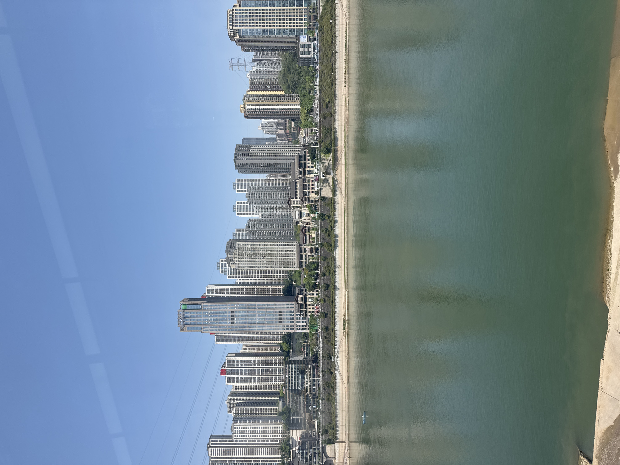 The image shows a waterfront cityscape in Yuzhong District, China, taken from an elevated perspective. A row of high-rise buildings lines the coast, varying in height, color, and architectural style. A narrow strip of green vegetation and a road separate the buildings from the shoreline. The water appears calm and slightly green, and a small boat is visible. The sky is clear and blue.