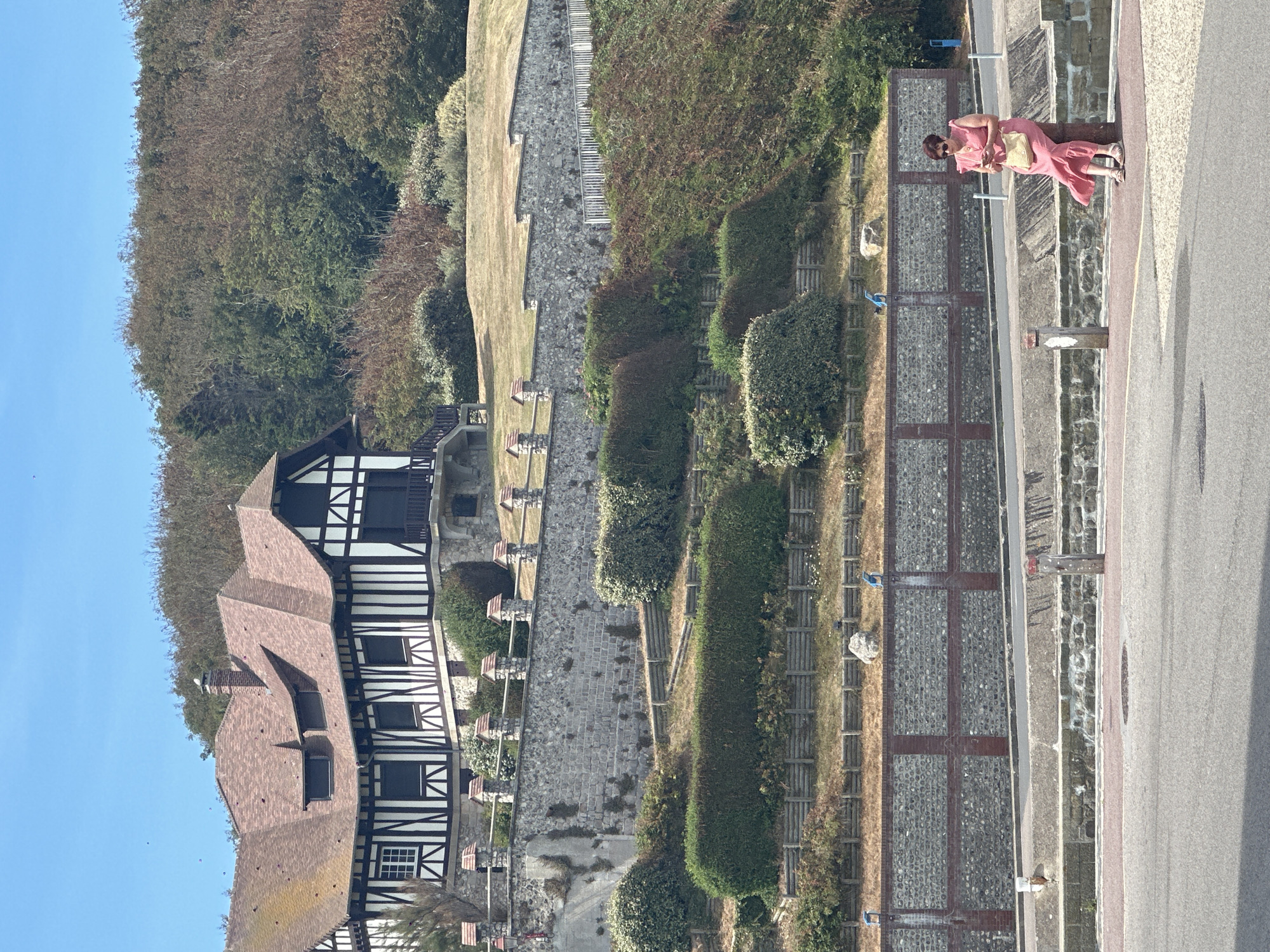A woman in a pink dress is sitting on a stone pillar near a road overlooking the coast in Saint-Valery-en-Caux, France. The woman is wearing sunglasses and holding a cell phone in her right hand. She has a straw purse slung over her left shoulder. The building behind her is a large house with a distinctive black and white half-timbered facade and a red tiled roof, situated on a hill. The terrain slopes down from the house, featuring stone walls, terraced gardens with neatly trimmed hedges, and wooden fencing. The sky is clear and blue.