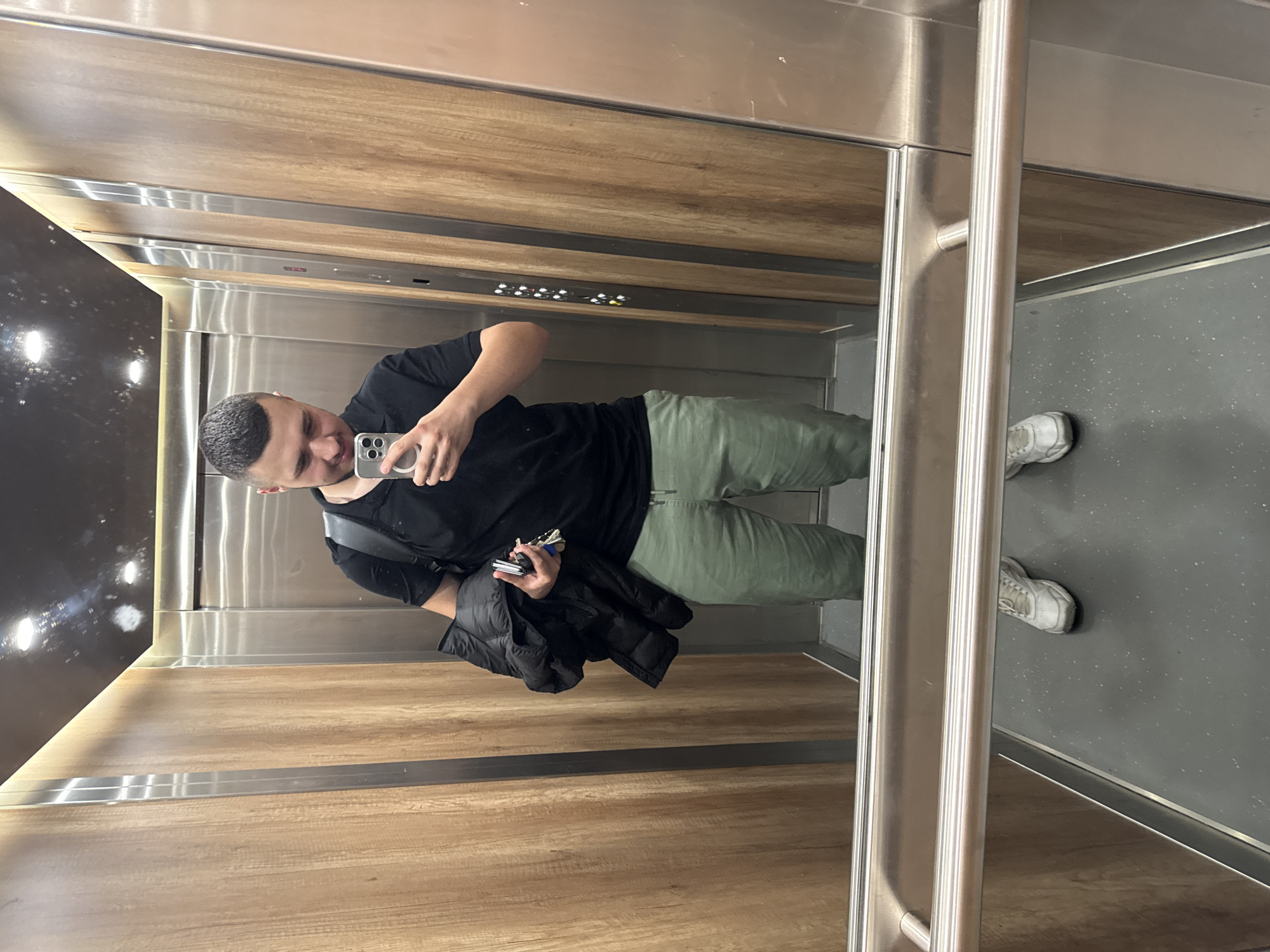 A young man takes a mirror selfie in an elevator with wood-paneled walls. He is wearing a black t-shirt, green pants, and white sneakers. He is holding a black jacket and keys in his left hand. The elevator has stainless steel accents and a handrail. The floor is grey with small flecks.
