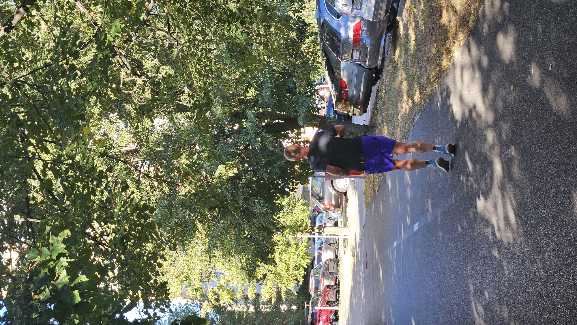 A man is jogging on a paved path lined with trees and parked cars. He is wearing a black t-shirt and blue shorts, with blue and white sneakers. The path is in a city, with buildings visible in the background. The man appears to be in the middle of his run, with sunlight filtering through the leaves of the trees, casting shadows on the path. The scene suggests a pleasant day for outdoor exercise in an urban environment. The location is identified as Prešov, Slovakia.