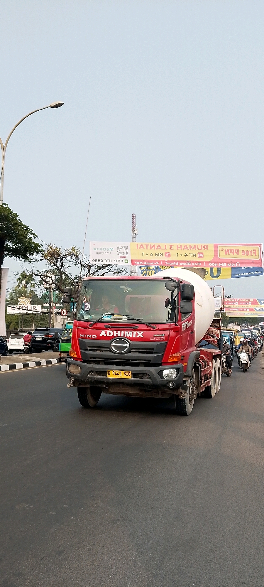 This is a vertical, eye-level, medium shot of a red HINO ADHIMIX cement truck in traffic on a sunny day in Gunung Putri, Indonesia. The truck is the main subject, occupying the majority of the frame and is positioned in the center. It is a large vehicle with a rotating drum for carrying cement. The front of the truck is detailed, showing the headlights, grille with the HINO logo, and the license plate "B 9441 SIO". A person is visible in the driver's seat.
The truck is moving forward on a paved road, indicated by the tire marks and the traffic in front of it. There are other vehicles on the road, including cars, motorcycles, and another truck. A line of motorcycles and scooters is visible behind the cement truck, forming a queue.
The environment suggests an urban or suburban setting. In the background, there are buildings and billboards. One billboard, with yellow and red text, is partially visible at the top right. There are also trees and a street lamp on the left side of the frame. The sky is bright and clear, indicating fair weather. The overall scene depicts a typical traffic situation in an Indonesian city.