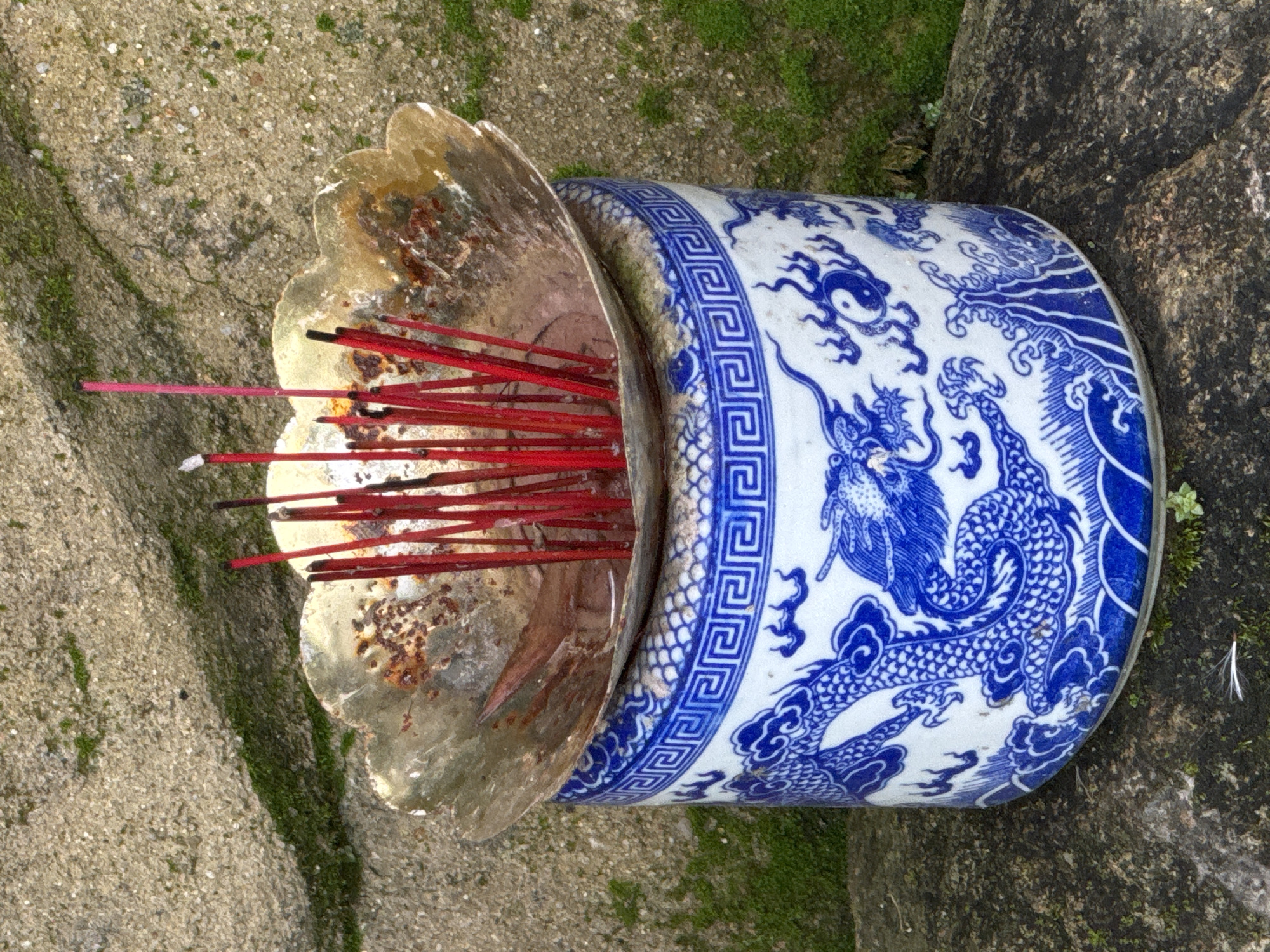 A close-up, outdoor, eye-level shot shows an incense burner with red incense sticks in it, placed on a weathered stone surface in what appears to be a garden or a place of worship. The incense burner consists of a blue and white ceramic pot with a dragon motif and a decorative blue band at the top, holding a tarnished, gold-colored, petal-shaped bowl filled with several red incense sticks. Some of the incense sticks are burning, with white smoke rising from them, and ash is visible in the bowl. The background is a textured, light brown stone wall with patches of green moss, suggesting an aged and possibly damp environment. The scene is likely located in Lâm Viên - Đà Lạt, Vietnam, given the style of the incense burner and the general aesthetic of such cultural sites in Vietnam. No people are present in the image.