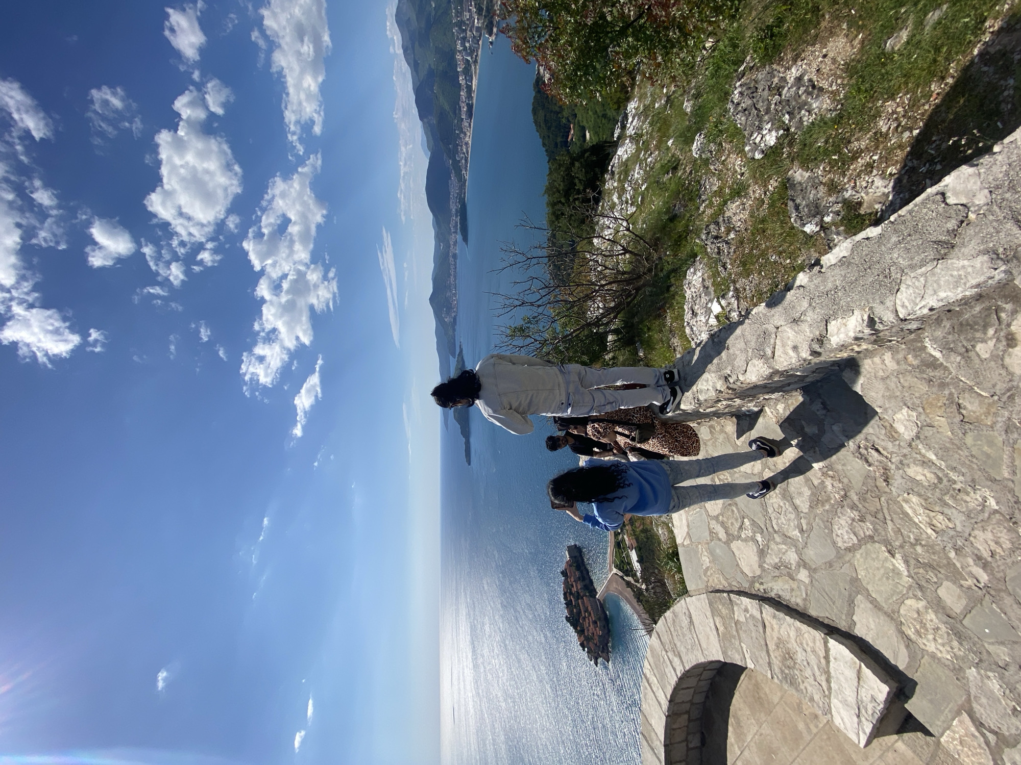 The image captures a panoramic view of a coastal landscape from a high vantage point, consistent with Sveti Stefan, Montenegro. The scene is dominated by a vast expanse of deep blue sea under a clear sky dotted with white cumulus clouds. Sunlight intensely reflects off the water's surface, creating a shimmering effect that extends towards the horizon.
In the middle ground, a distinctive island or peninsula, densely covered with terracotta-roofed buildings, is prominently featured, connected to the mainland by a narrow causeway. Along the coastline in the distance, a town with numerous buildings is visible, nestled against verdant mountains that rise steeply from the sea.
In the foreground, a stone-paved viewing platform or path provides a sturdy base. Three individuals are standing on this platform, their backs mostly to the camera, absorbed in the breathtaking scenery. One person, wearing a light-colored jacket and trousers, stands slightly to the left, gazing out. To their right, another individual in a blue top and light trousers holds a smartphone, seemingly capturing the view. A third person, partially visible behind them, wears dark attire and carries a leopard-print bag.
The time of day appears to be bright daytime, indicated by the strong sunlight and reflections. The weather is clear and sunny. No visible text is present in the image. The overall impression is one of serene admiration for the beautiful natural and architectural landscape.