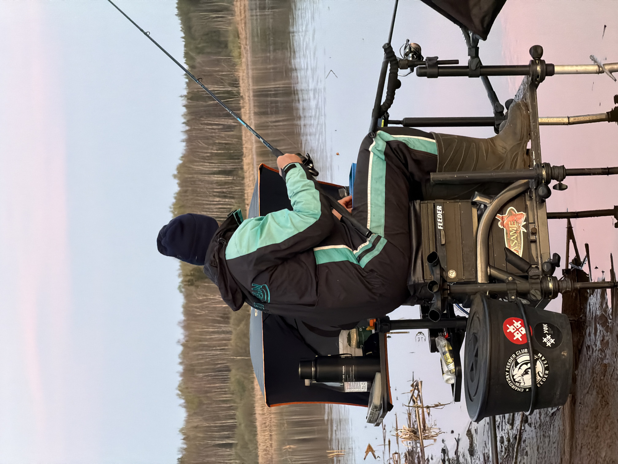 A person is fishing from a specialized chair at a lake. The person wears a dark jacket with light blue accents, dark pants with a light blue stripe, and dark boots. They are holding a fishing rod, and a dark blue hat is visible. The fishing chair has various attachments and storage containers. One of the containers has a sticker with the text "CHAIKOVSKY FEEDER CLUB TEAM". Reeds and reflective water are visible in the background, along with a treeline. The sky is pale.