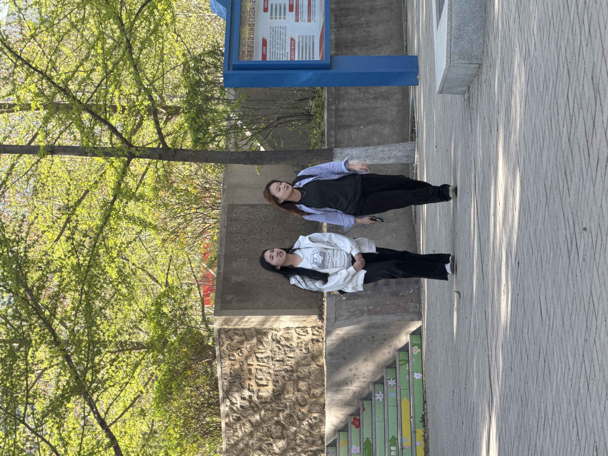 Two young women are standing on a sidewalk next to a retaining wall and a sign in Zhongtun, China. The woman on the right is wearing a black t-shirt and pants with a light blue and white striped shirt over the top. The woman on the left is wearing a white t-shirt and black pants with a white hoodie over the top. A tree with green leaves is to the left.
