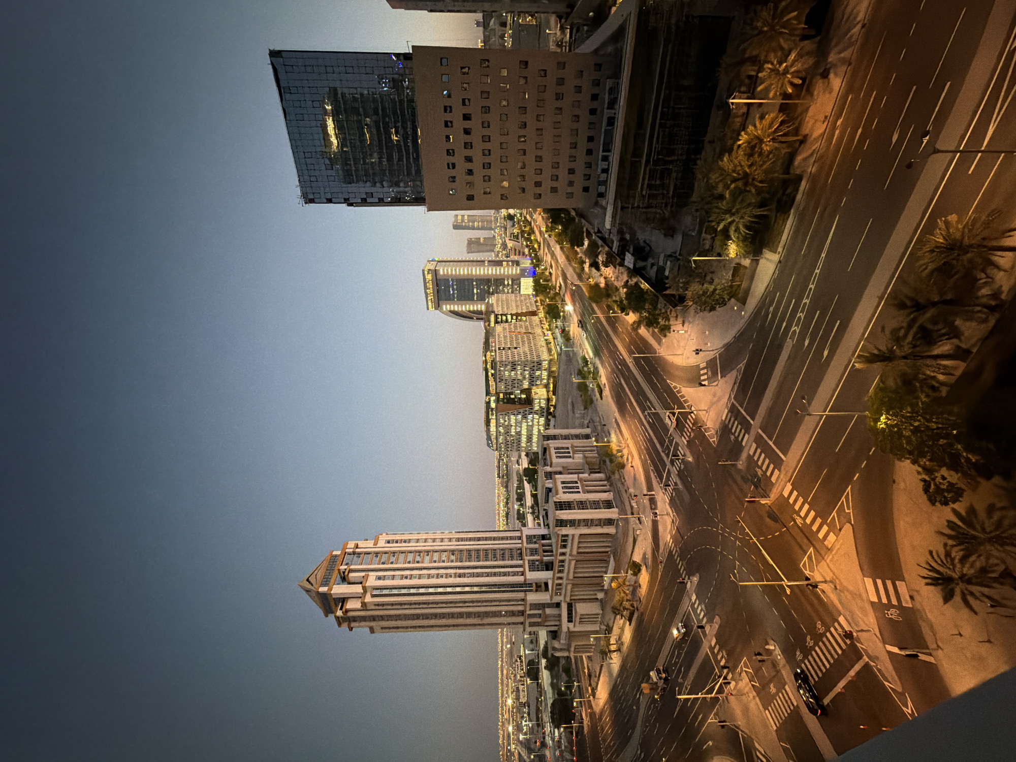 This is a high-angle, full-shot, eye-level, medium, long, and wide view of a city intersection at dusk. The sky is a gradient of deep blue to a lighter gray, indicating twilight. Several modern buildings are visible, including a tall, beige skyscraper with a stepped roof in the left foreground, a square, dark glass skyscraper to the right, and a cluster of illuminated, modern office buildings in the background. The intersection is well-lit by streetlights and the interior lights of the buildings, casting warm orange and yellow glows. Traffic lines are clearly marked on the asphalt roads, and pedestrian crossings are visible. A few vehicles, including a truck and a car, are present on the roads, along with a few individuals walking. Palm trees are scattered throughout the scene, adding a tropical element to the urban landscape. The overall impression is a vibrant, active city preparing for the night.