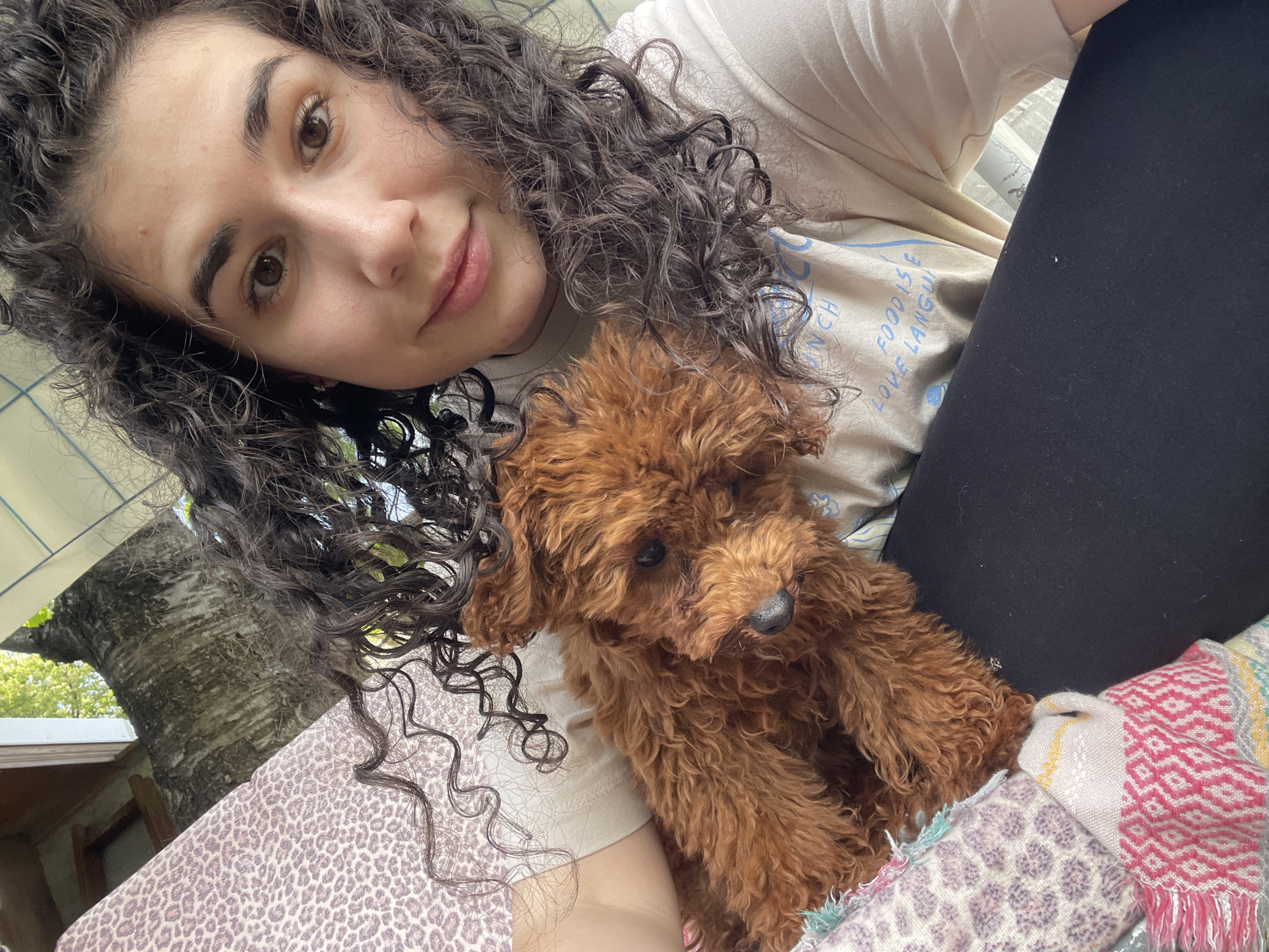 A selfie of a young woman with curly hair and a small, brown poodle-like dog. The woman is fair-skinned with brown eyes and eyebrows. She's wearing a beige t-shirt with blue writing and black pants. The dog is sitting on her lap. Behind them is a mottled background of leaves, tree bark, and a white roof. The location can not be verified as Sofia, Bulgaria.