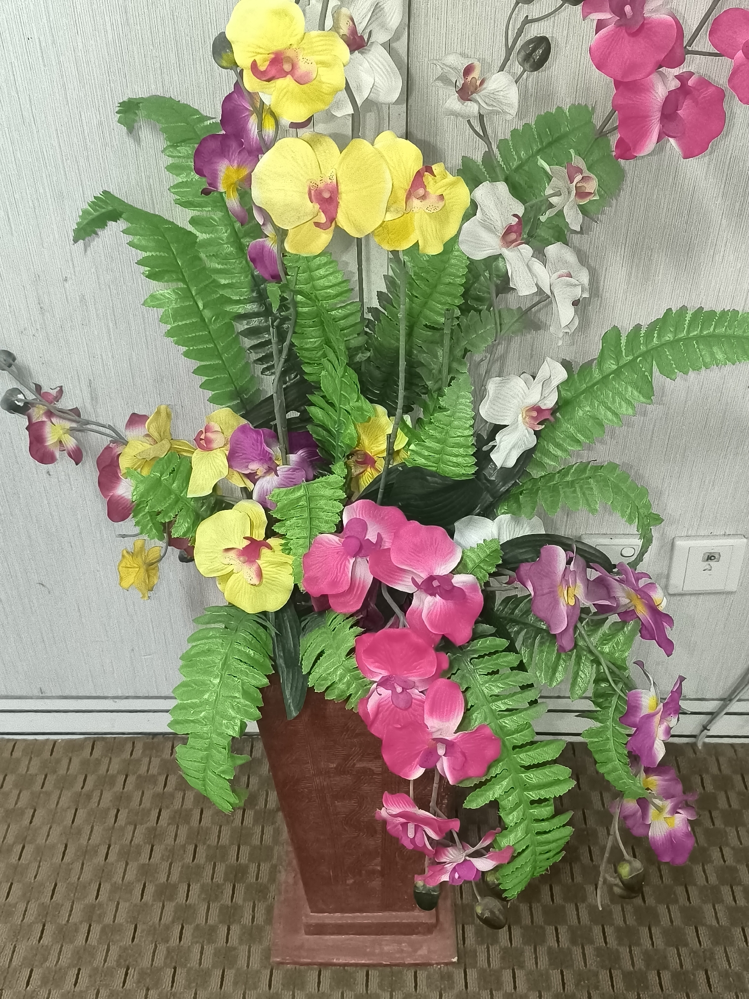 A tall, artificial floral arrangement of orchids and ferns is displayed in a brown vase. The orchids are in various shades of yellow, pink, and white, with some featuring deep pink accents. The fern leaves are a vibrant green. The arrangement is positioned on a textured, brown mat against a light-colored, vertical-lined wall. To the right of the arrangement, a white electrical outlet is visible on the wall. The image appears to be taken indoors, possibly in a residential or commercial setting in Kalabbirang, Indonesia. There are no people or animals present in the scene.