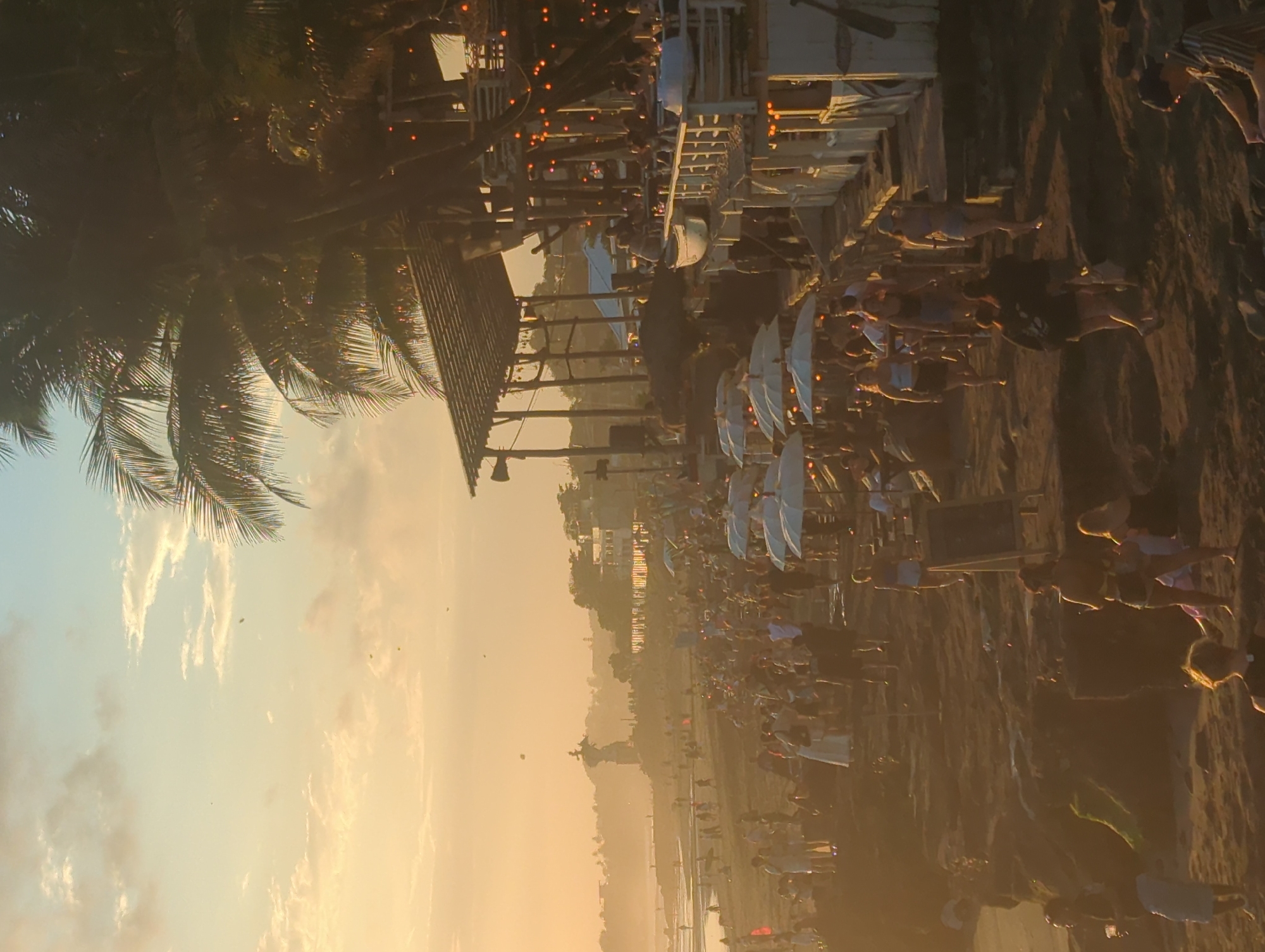 The image depicts a beach scene at sunset in North Kuta, Indonesia. The beach is crowded with people, many of whom are seated at tables under white umbrellas. In the foreground, there are palm trees and a wooden structure, possibly a beach bar or restaurant, with lights strung across it. The setting sun casts a golden light over the entire scene, creating a hazy atmosphere. The ocean is visible in the background, with waves breaking on the shore. The image appears to be taken from a slightly elevated position, looking out over the beach towards the horizon.