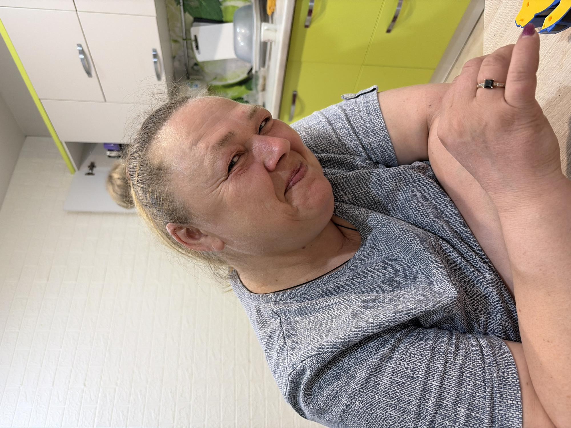 A woman is indoors, in what appears to be a kitchen, based on the cabinetry in the background. She is wearing a grey shirt and a ring with a dark stone. The location is not discernible based on the details provided.
