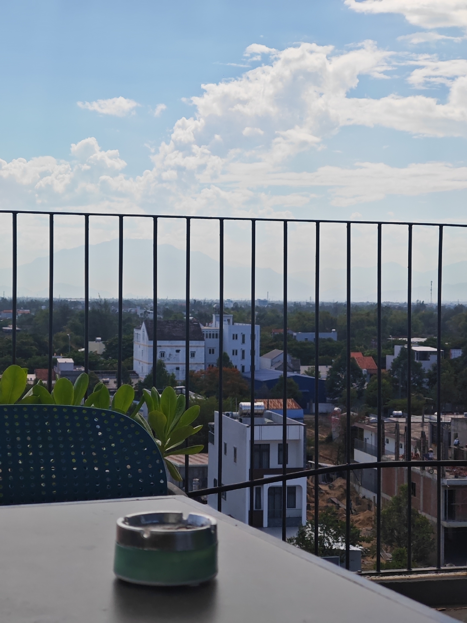 A clear sky with scattered white clouds looms over a cityscape in Đà Nẵng, Vietnam. In the foreground, a metal railing with vertical bars separates the viewer from the scene beyond. A dark green chair with a perforated seat is visible to the left, and an ashtray sits on a gray table in the lower right corner. The cityscape features a variety of buildings, including residential structures, some appearing to be under construction, with exposed brickwork and scaffolding. Lush green trees dot the landscape, interspersed with the architecture. In the distance, hazy mountains form a backdrop against the horizon. The overall impression is a panoramic view from a rooftop or balcony, overlooking a developing urban area on a sunny day.